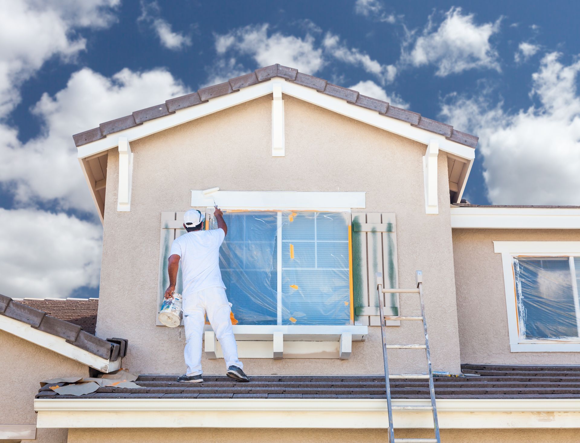 Un ouvrier peint le mur extérieur d'une maison sur une échelle sous un ciel bleu.
