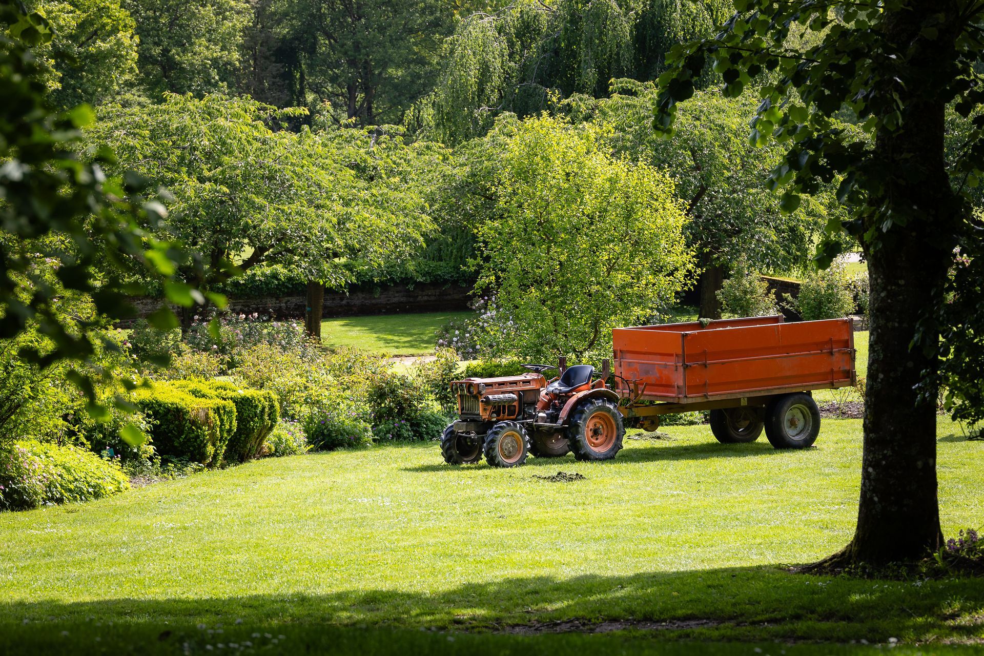 Tracteur orange avec remorque dans un jardin bien tondu entouré d'arbres.