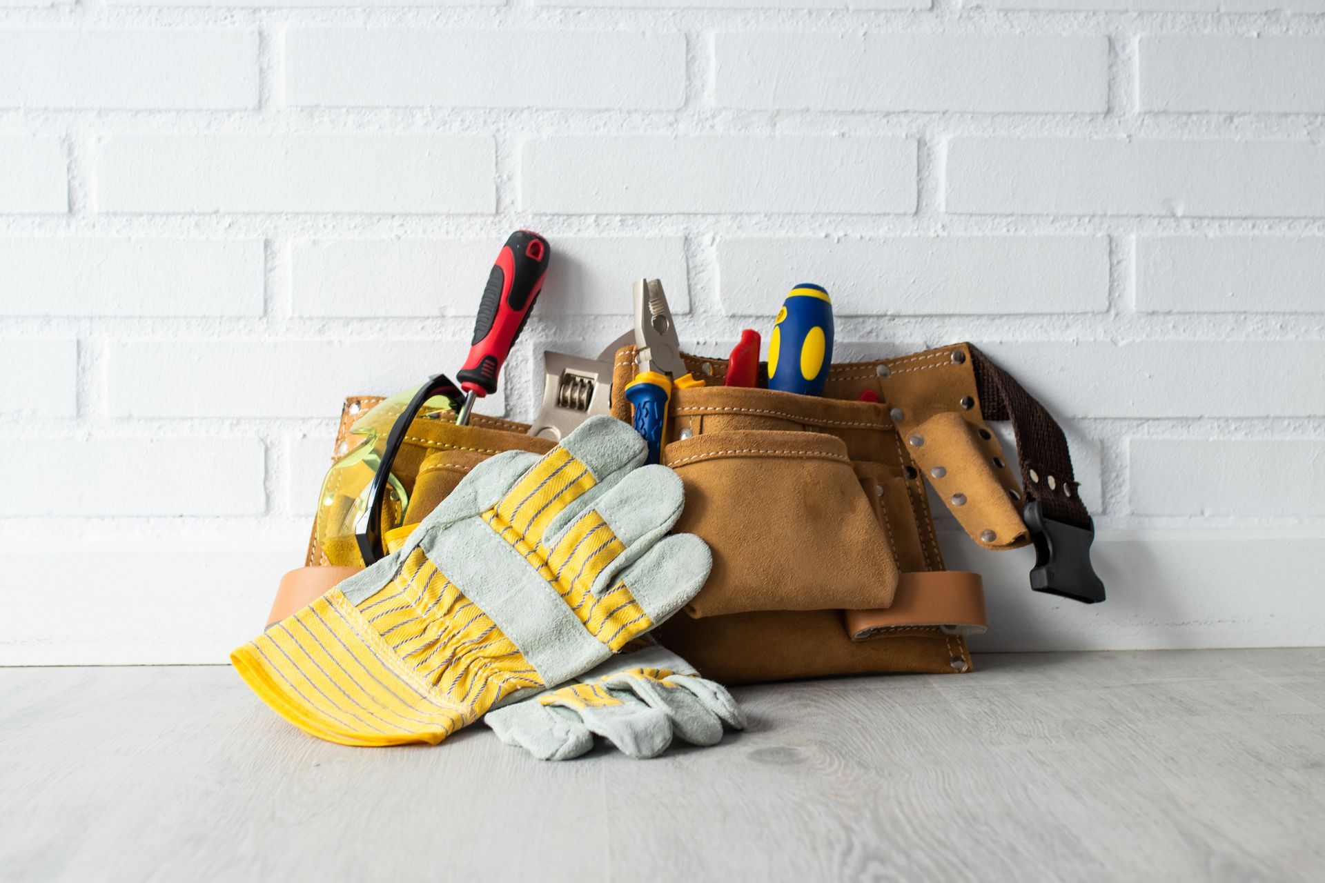 Gants de travail en cuir et outils à main dans une ceinture à outils sur une table blanche contre un mur de briques