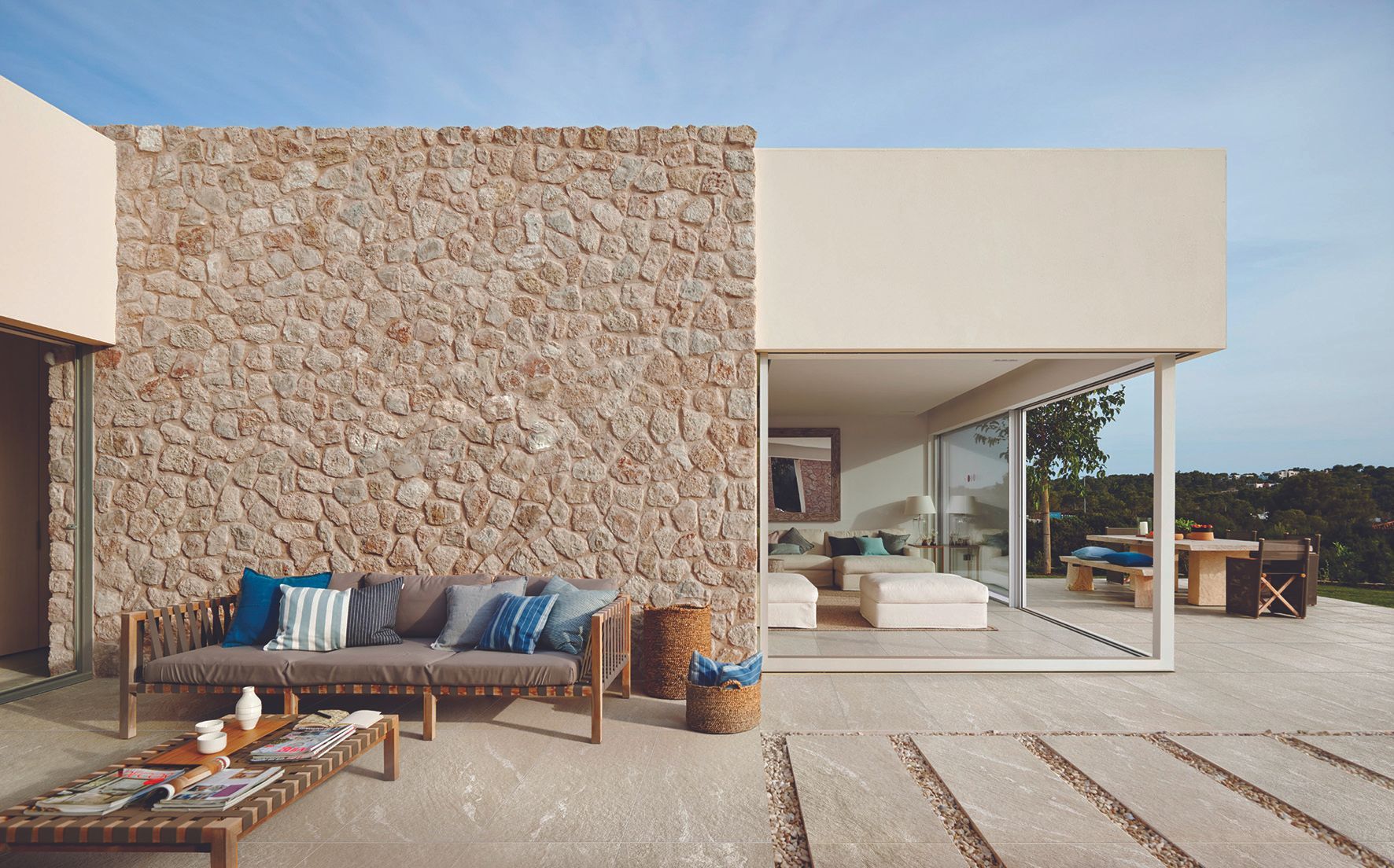Patio moderne avec mur en pierre, canapé d'extérieur et portes vitrées donnant sur une maison lumineuse et minimaliste
