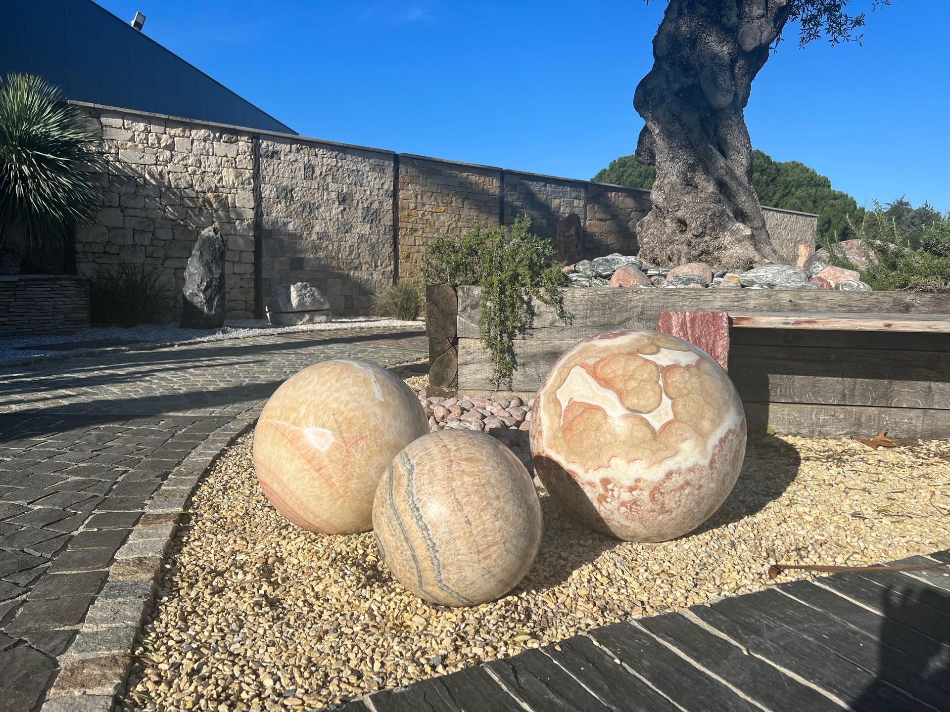 Trois grandes sphères de pierre sur un chemin de gravier, près d'un mur de pierre et d'un arbre sculpté sous un ciel bleu.