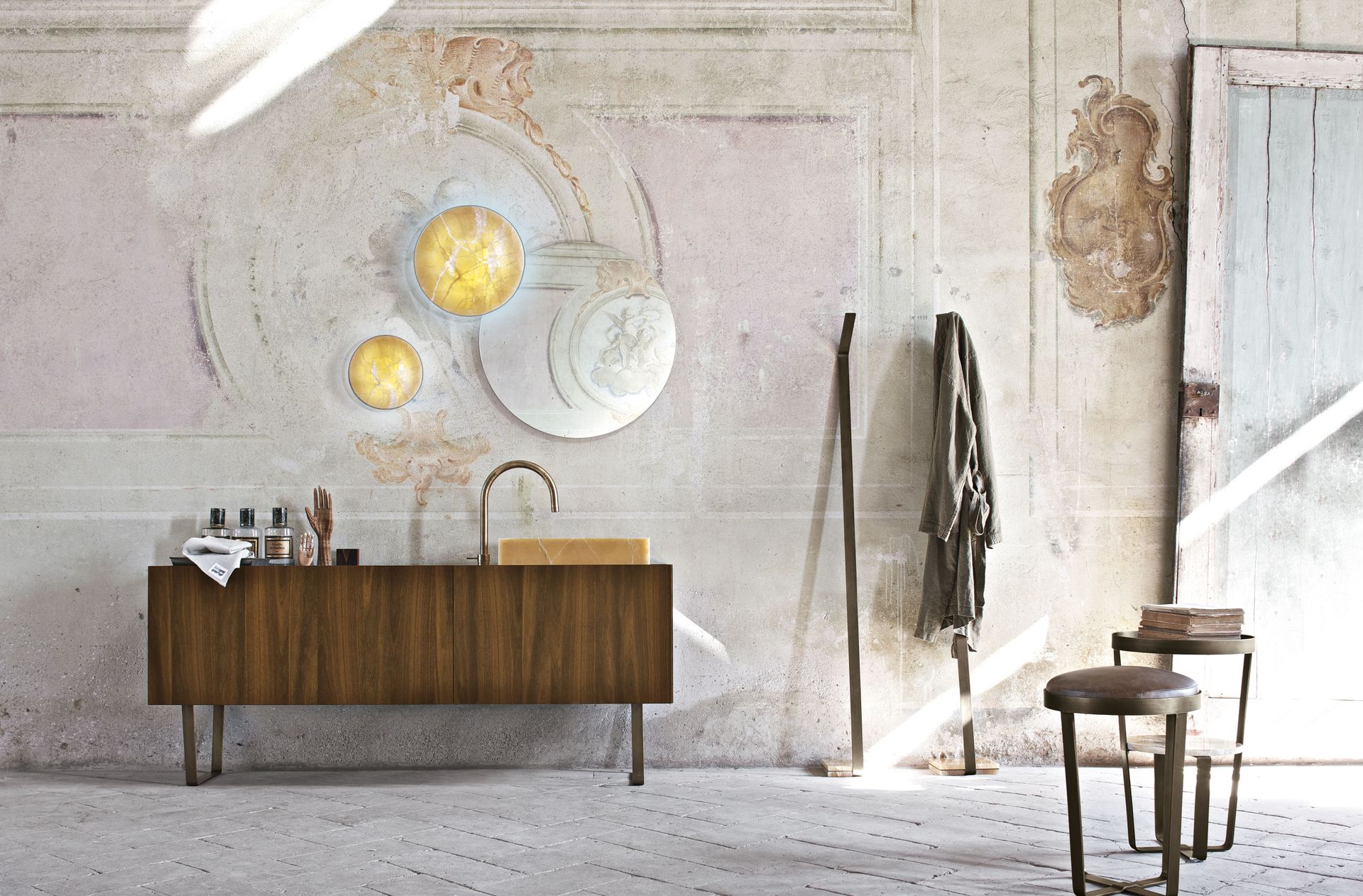 Chambre minimaliste avec un buffet en bois, des tableaux, des manteaux suspendus et une chaise près d'un mur ensoleillé.