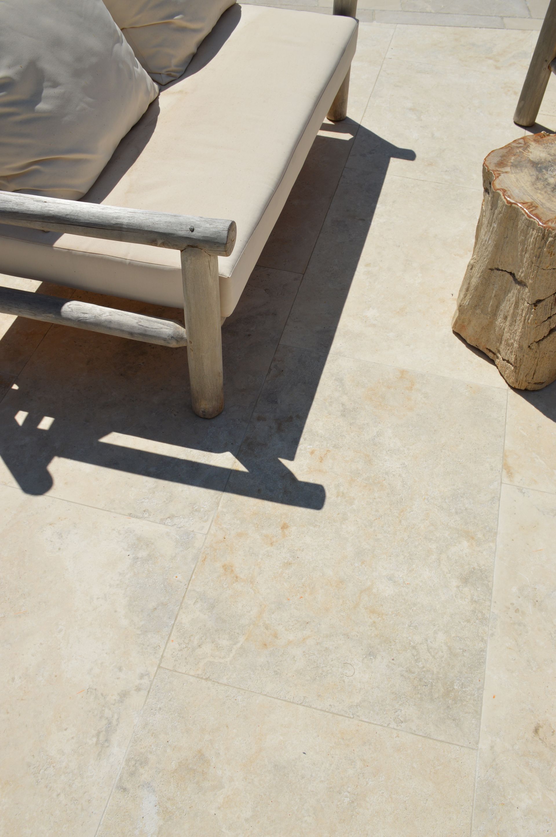 Terrasse extérieure avec banc beige rembourré, coussin et table en pierre sur un dallage ensoleillé.