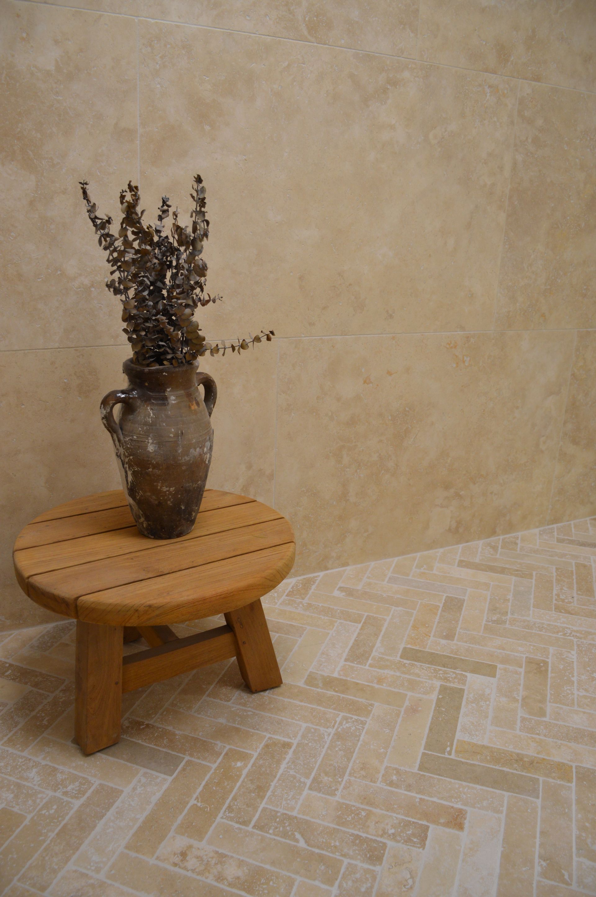 Vase de fleurs séchées posé sur un tabouret rond en bois, devant un mur beige et un sol carrelé.