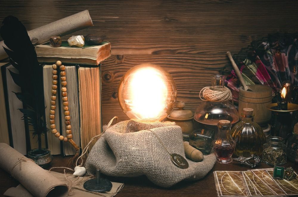 Bola de cristal brillante en un soporte de piedra, rodeada de libros, pergaminos, pociones y velas sobre una mesa de madera.