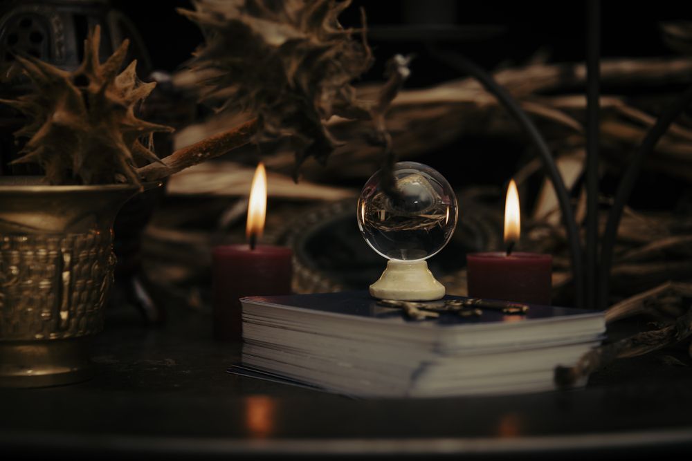 Bola de cristal sobre libro, flanqueada por velas, con follaje seco, sobre una mesa oscura.