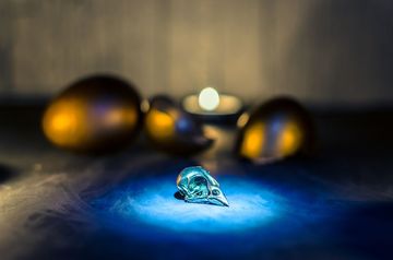 Un cráneo de pájaro de cristal iluminado en azul reposa entre el humo, con tres huevos dorados y una vela en el fondo.
