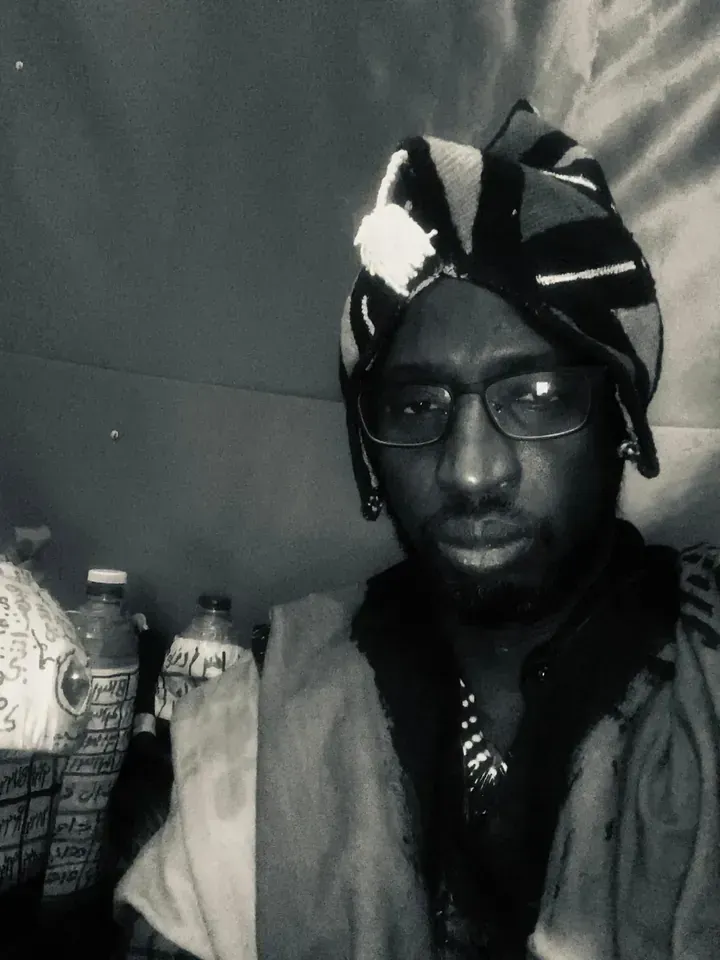 Persona con gafas y pañuelo estampado en la cabeza, mirando a la cámara. Se ven botellas al fondo.