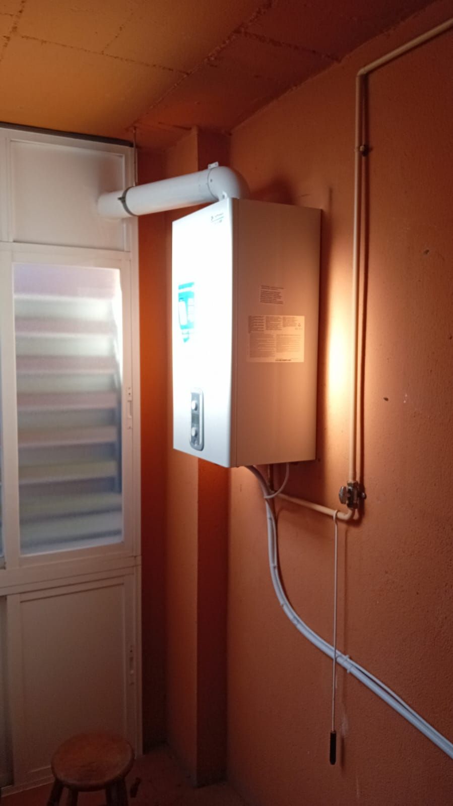 Calentador de agua blanco sobre pared naranja con tubo de ventilación que se extiende hacia el exterior.