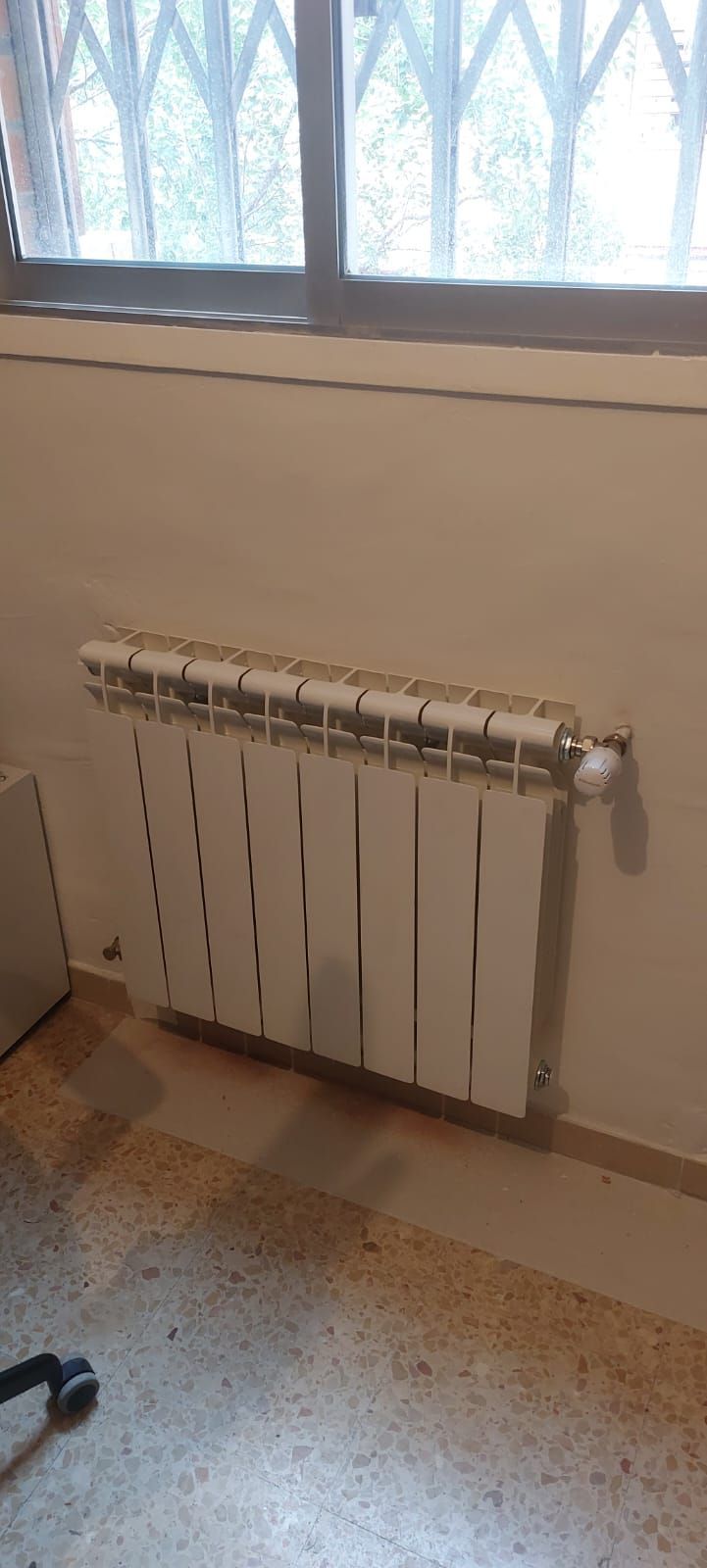 Radiador blanco montado en una pared beige debajo de una ventana con reja de seguridad.