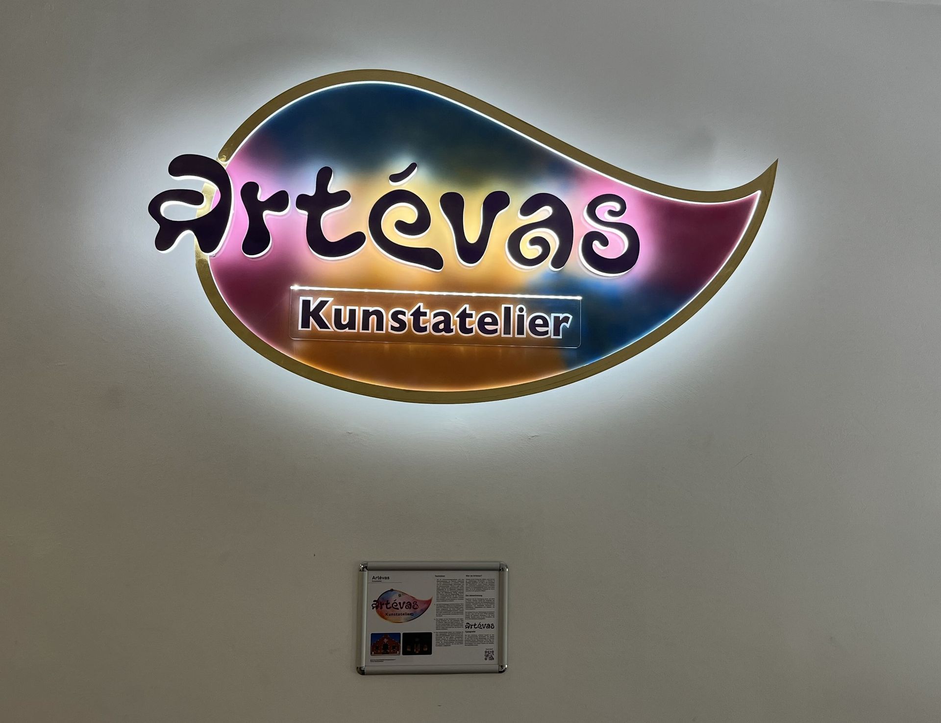 Beleuchtetes Artevās Kunstteller-Schild an einer Wand, leuchtend in Lila, Rosa und Gold.