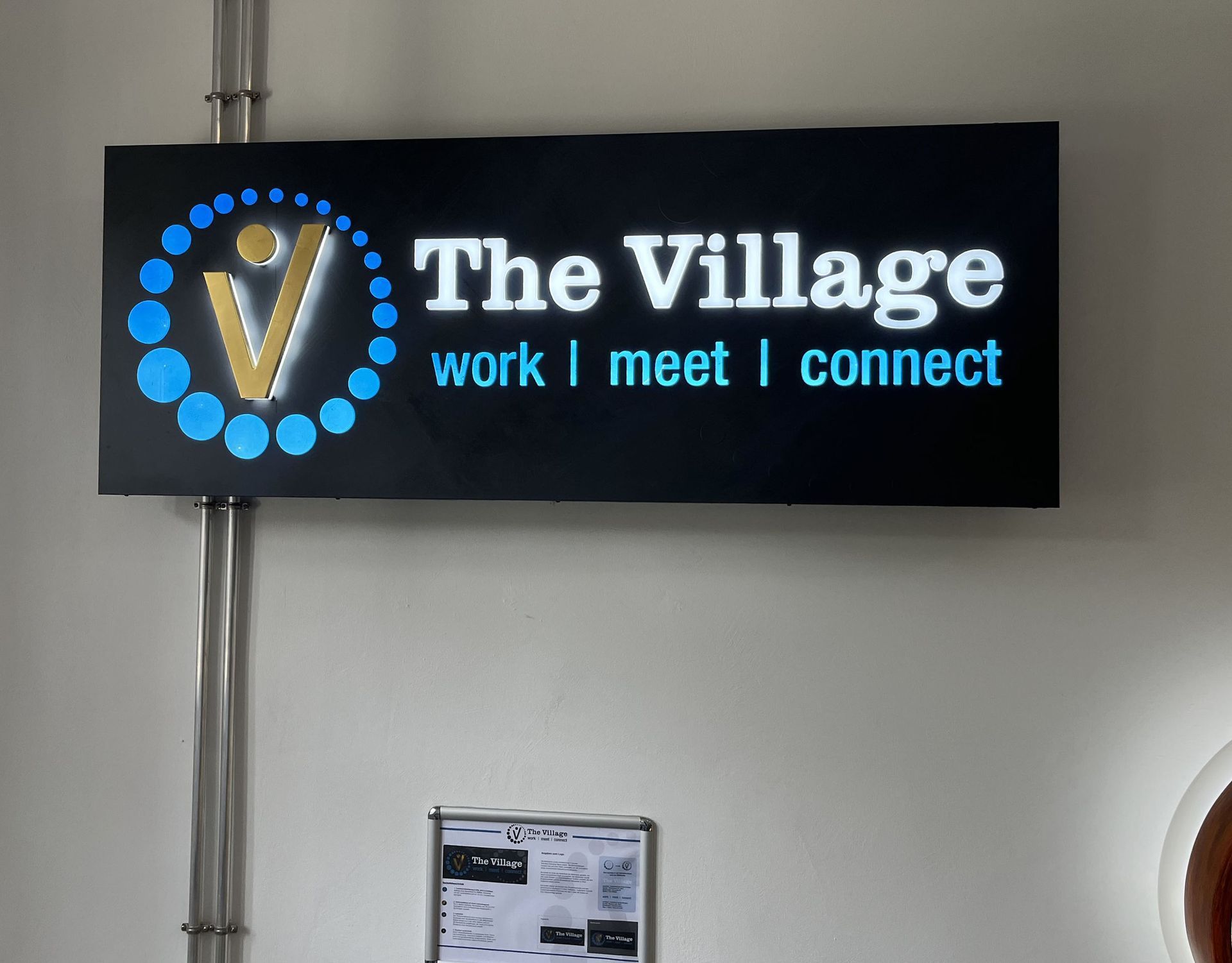 Wandschild für „The Village“ mit blauem Logo und dem Schriftzug „Arbeit | Treffen | Vernetzen“ auf schwarzem Hintergrund