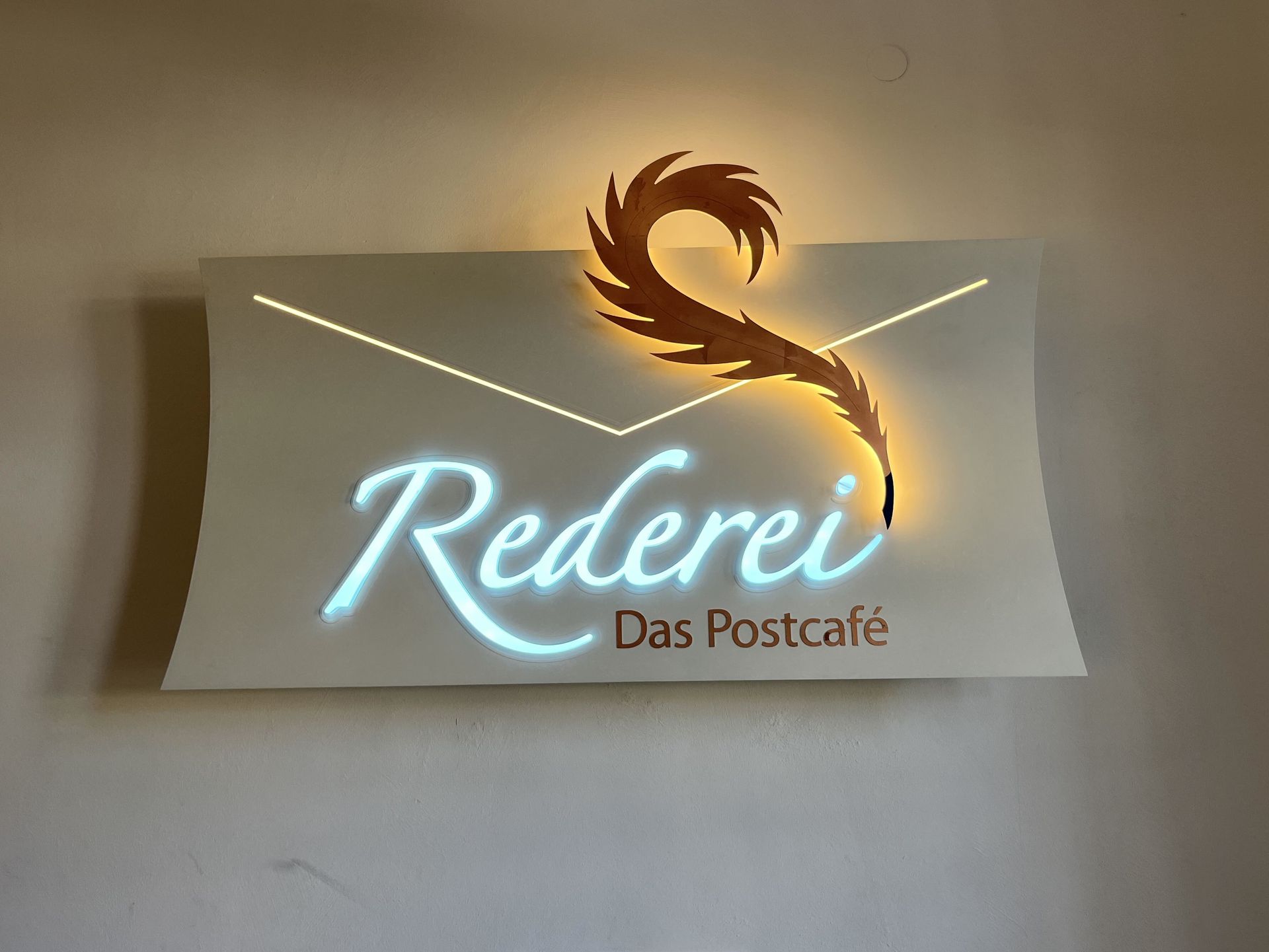 Ein Schild der Rederei Das Postcafé mit leuchtender Schrift und einem goldenen Vogellogo an einer Wand