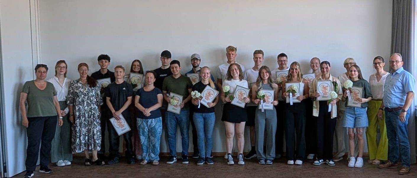 Eine Gruppe von Menschen steht in einem Raum, hält Papiere und Blumensträuße in den Händen.