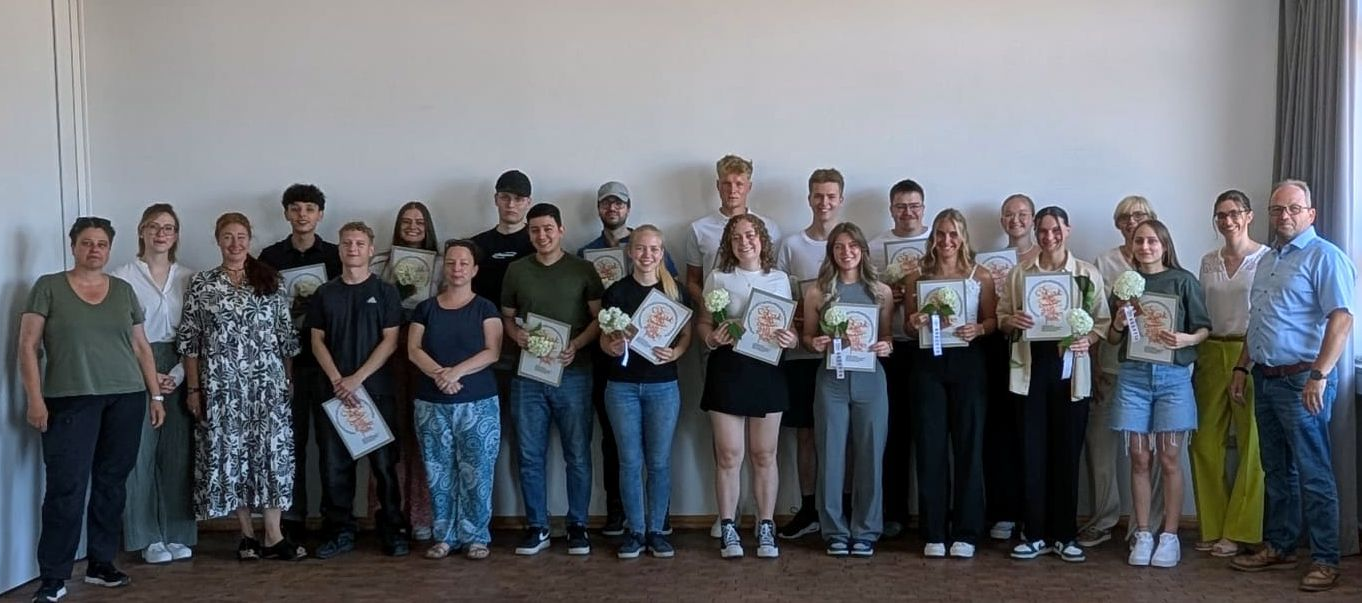 Eine Gruppe von Menschen steht in einem Raum, hält Papiere und Blumensträuße in den Händen, vermutlich eine Preisverleihung.