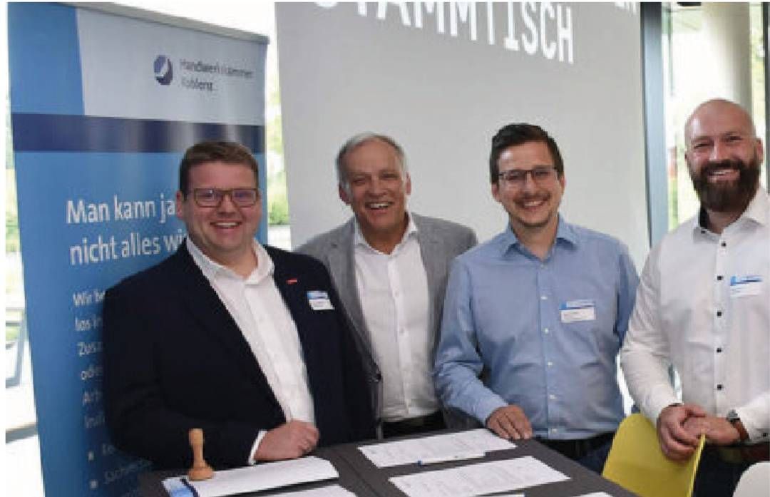 Vier lächelnde Männer an einem Tisch, bei einer Veranstaltung. Banner und offene Dokumente liegen vor.