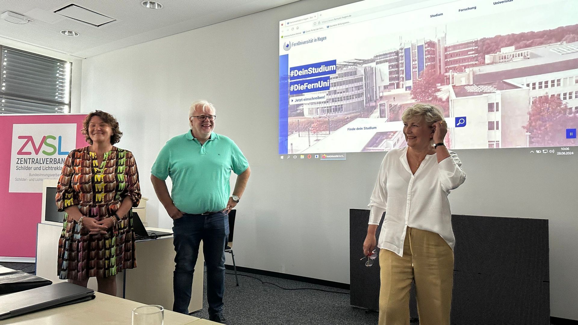 Ein Mann und zwei Frauen stehen in einem Konferenzraum vor einem großen Bildschirm.