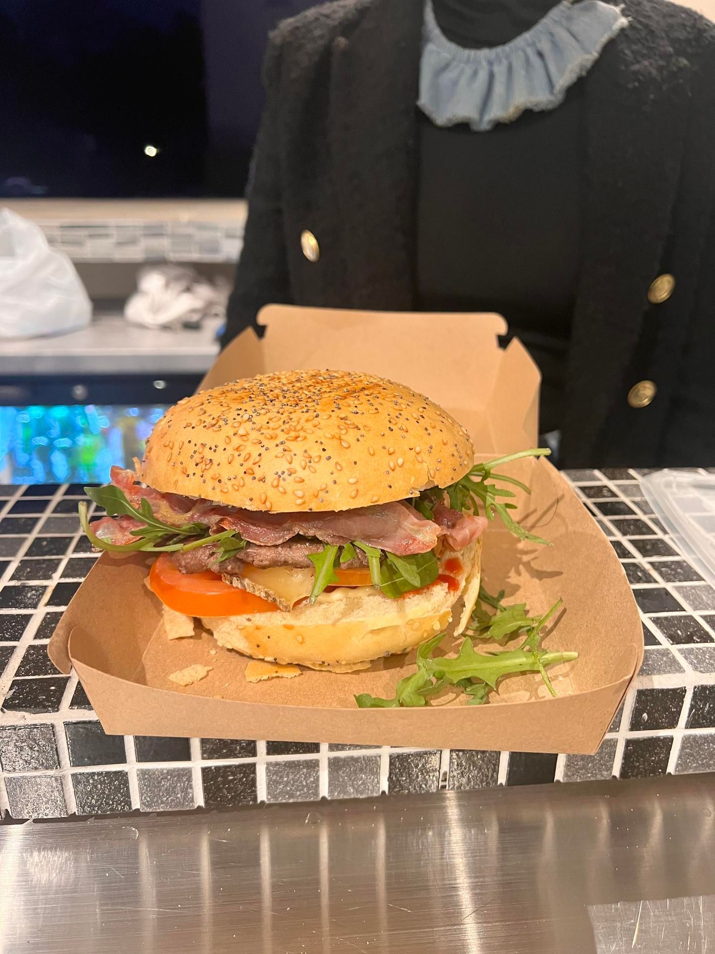 Un hamburger au sésame servi dans un plateau en carton sur un comptoir carrelé en noir et blanc, avec une personne derrière.