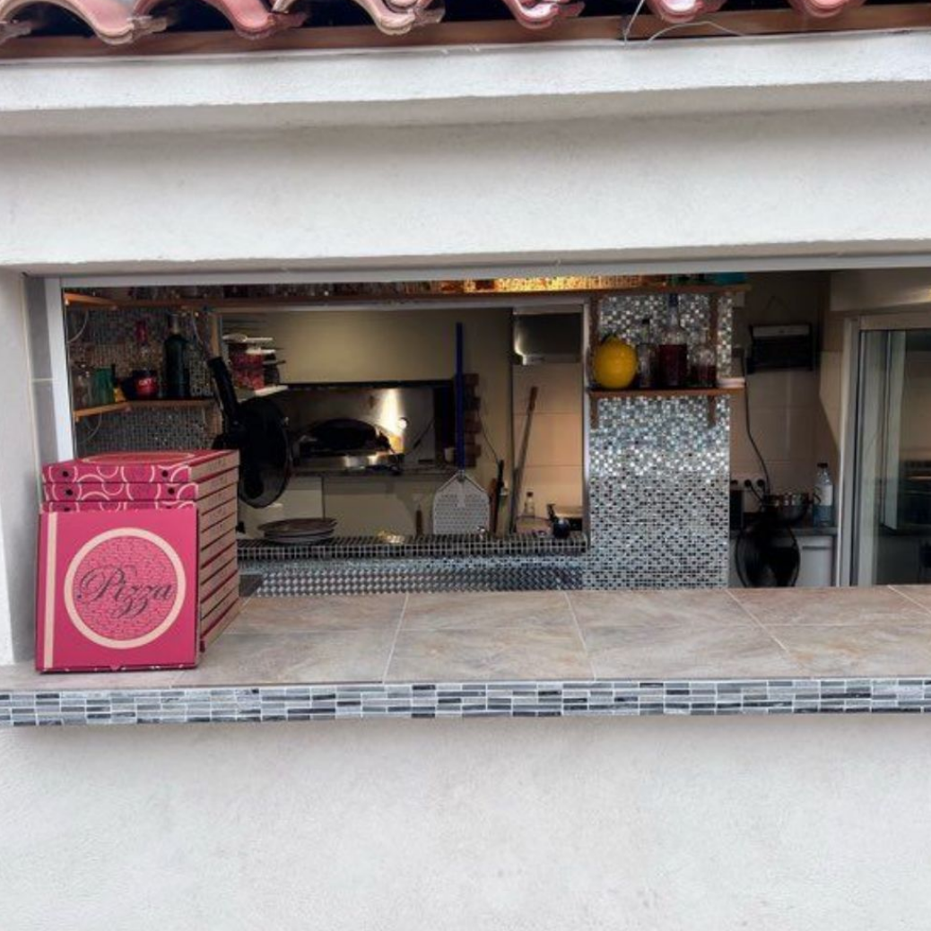 Fenêtre ouverte d'un café avec comptoir carrelé, enseigne rose et étagères à l'intérieur.