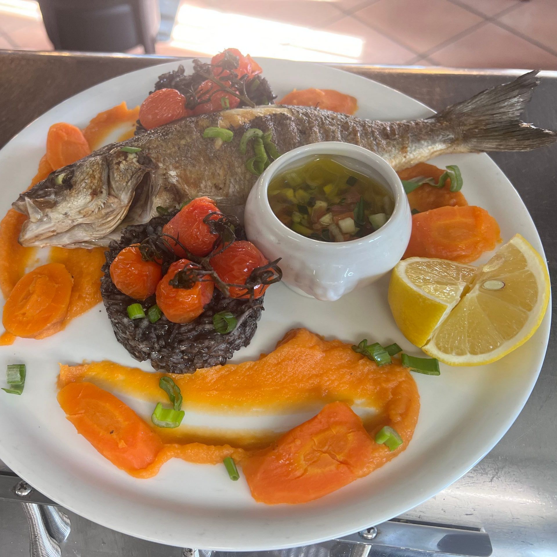 Poisson entier grillé servi sur une assiette blanche, accompagné de sauce à l'orange, de quartiers de citron et d'un petit bol de salsa.