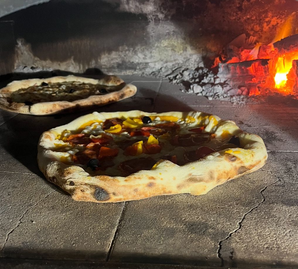Deux pizzas cuisent dans un four à bois, celle de devant avec sa garniture fondue et les flammes qui brillent à droite.