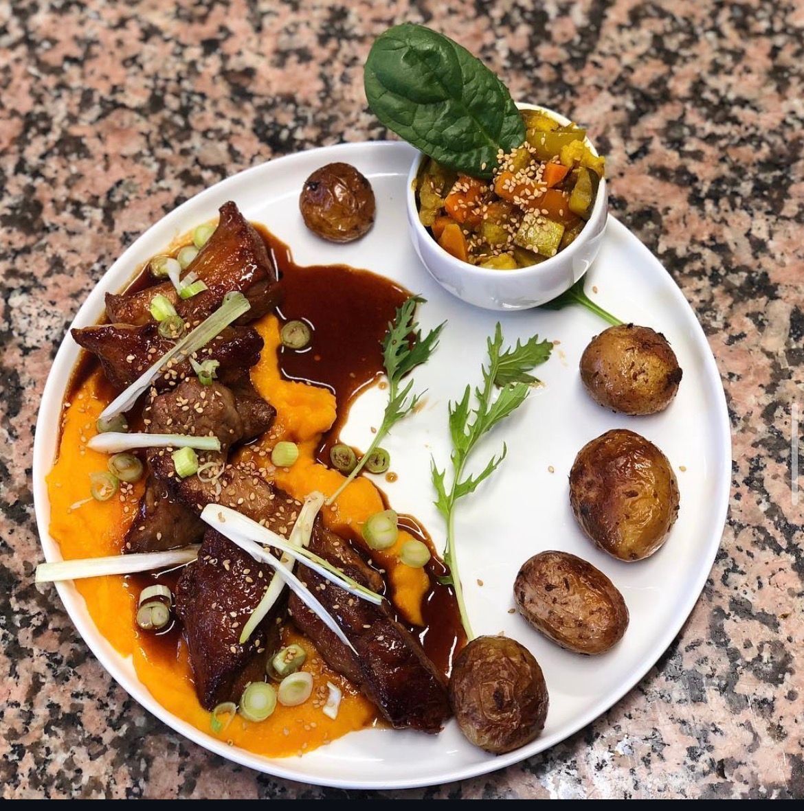 Plat gastronomique présenté à l'assiette, avec sauce brune, purée d'orange, oignons verts, câpres et une petite coupelle de garniture à côté.