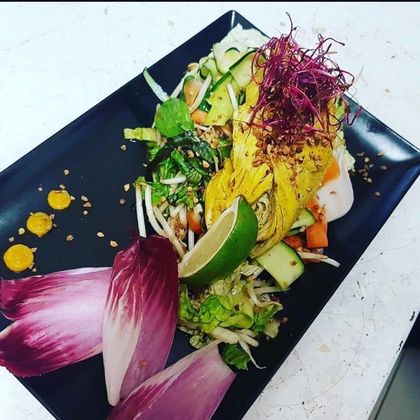 Salade colorée dressée sur une assiette rectangulaire noire, avec quartier de citron vert, radicchio violet, herbes et légumes.