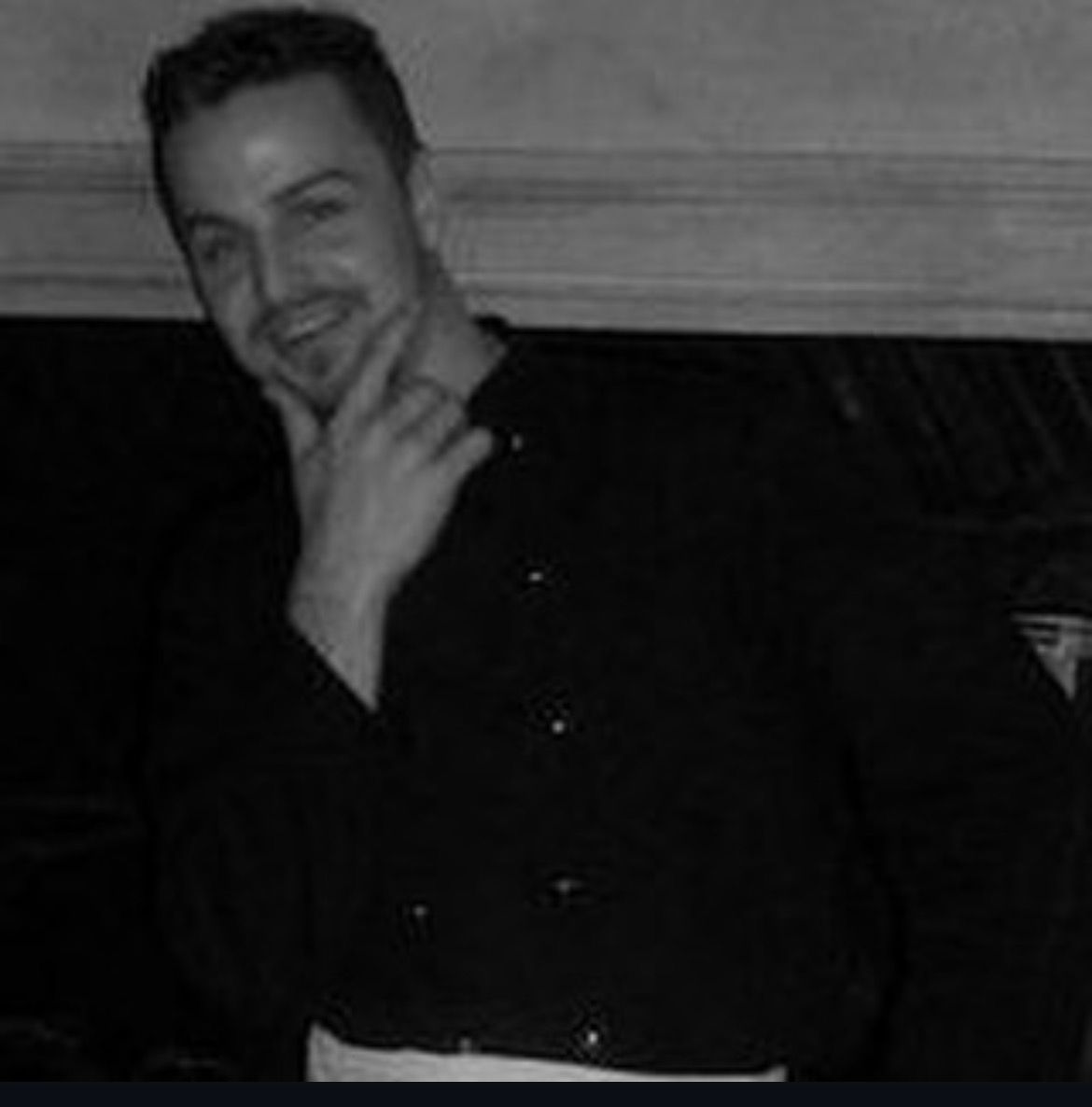 Un homme souriant, vêtu d'une chemise sombre, pose à l'intérieur, la main sur le menton.