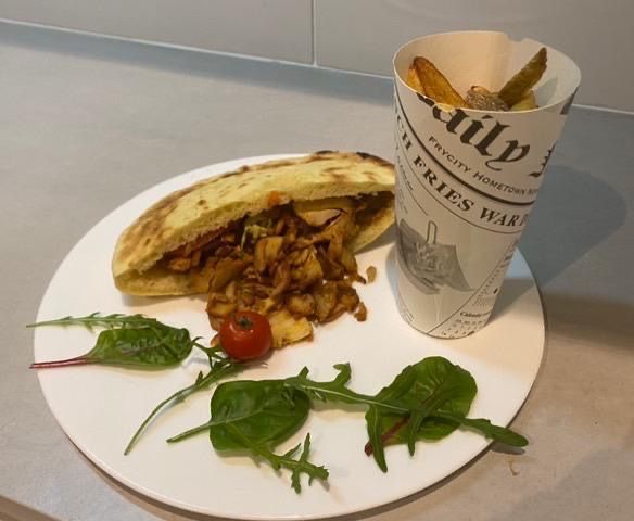 Sandwich grillé avec salade et tomate sur une assiette blanche, à côté d'un cornet de frites en papier sur une table.