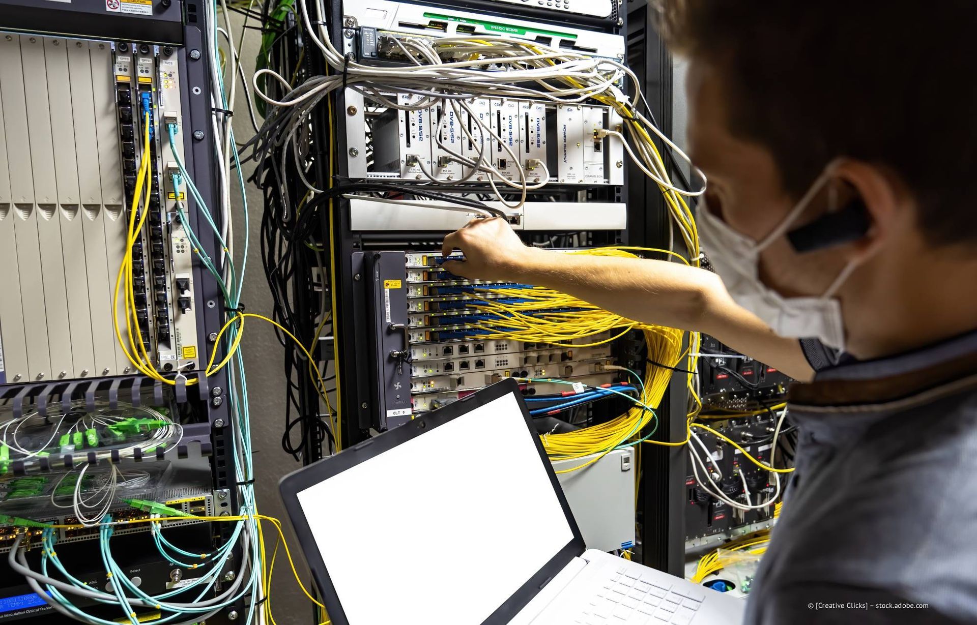 Techniker mit Laptop arbeitet an einem Netzwerkserver in einem Serverraum. Er trägt eine Gesichtsmaske.