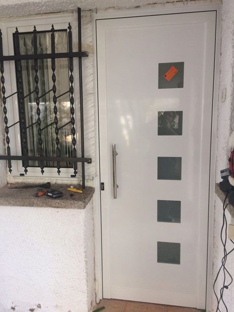 Una puerta blanca con cuadrados está al lado de una ventana con rejas.