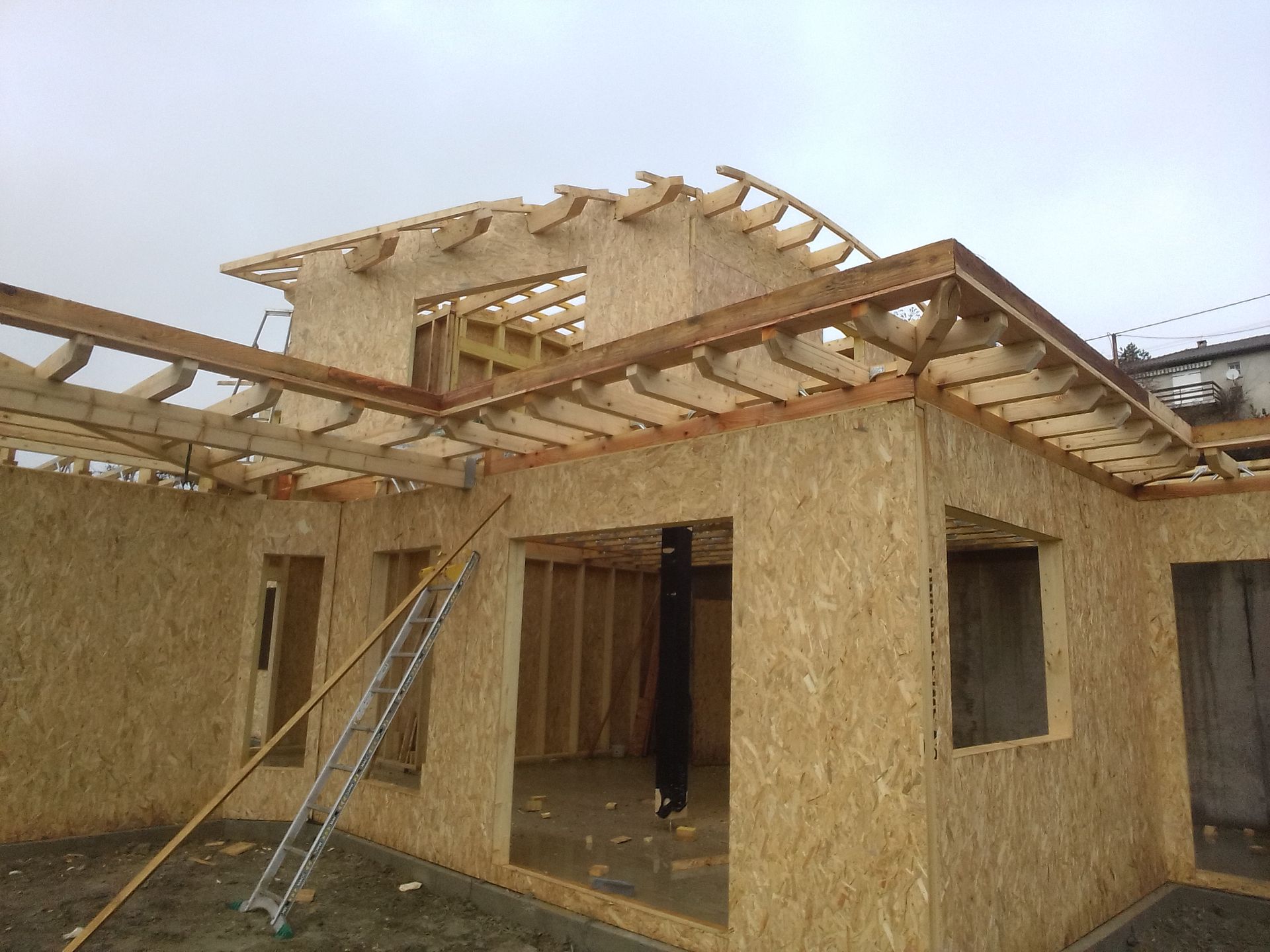 Maison en construction ; ossature en bois avec revêtement en panneaux OSB. Charpente du toit visible. Temps nuageux.
