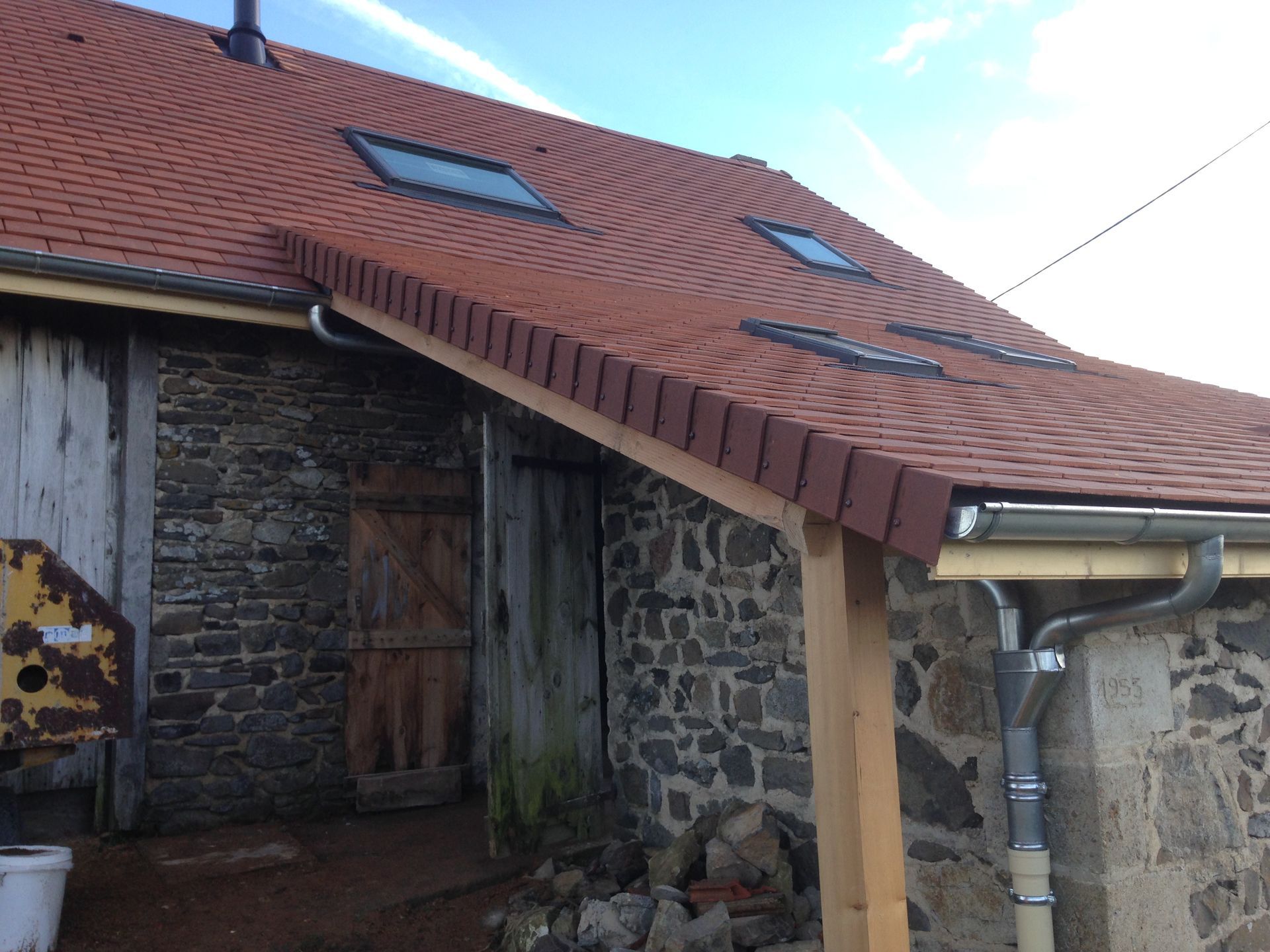 Maison en pierre avec toit de tuiles rouges, puits de lumière et auvent en bois.
