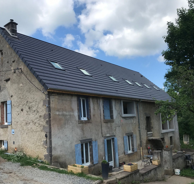 Maison en pierre de deux étages avec toit de tuiles sombres, plusieurs puits de lumière, volets bleus et ciel nuageux.