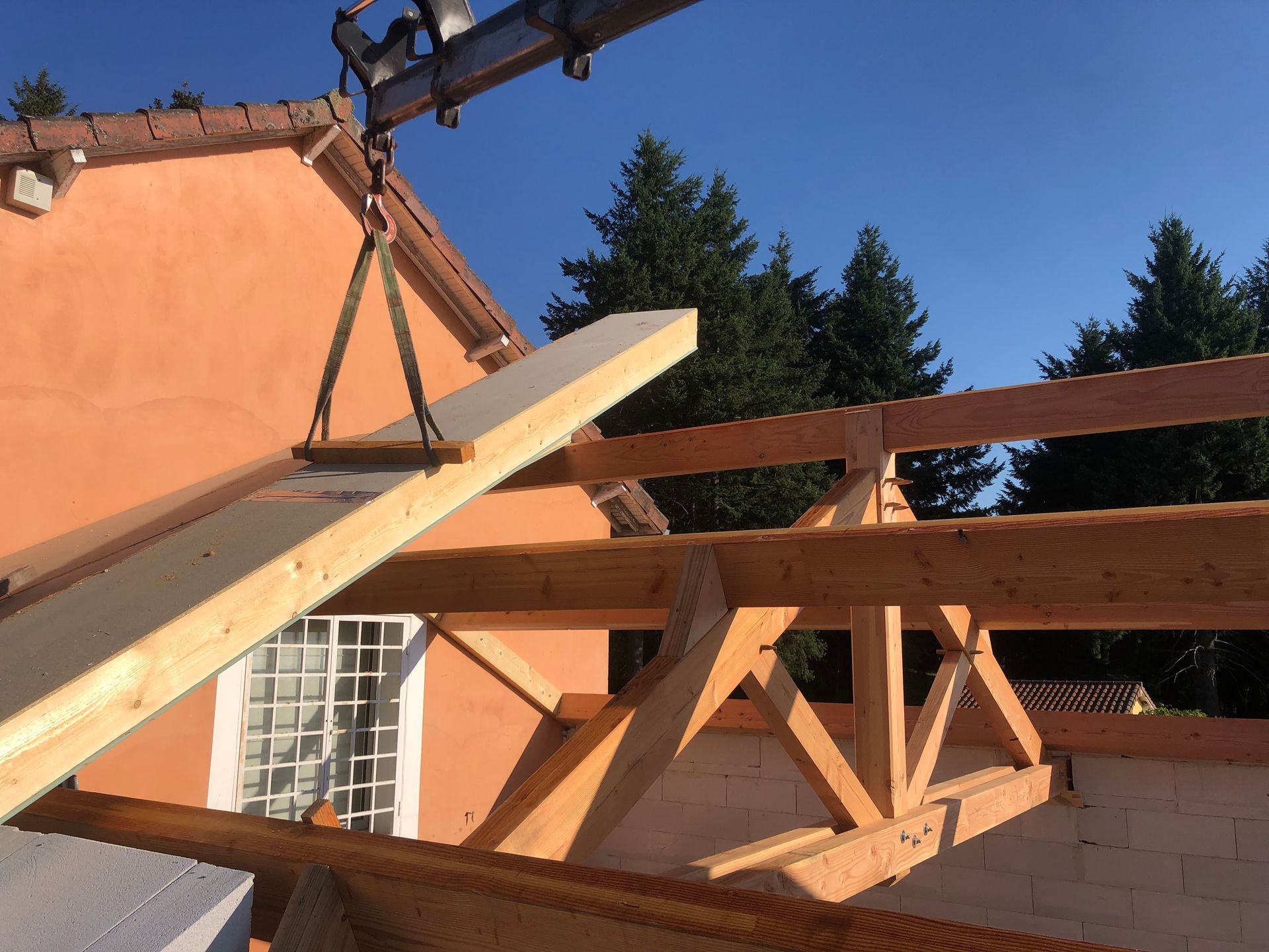 Grue soulevant une poutre en bois pour la poser sur la charpente d'une maison en construction. Maison orange, ciel bleu.