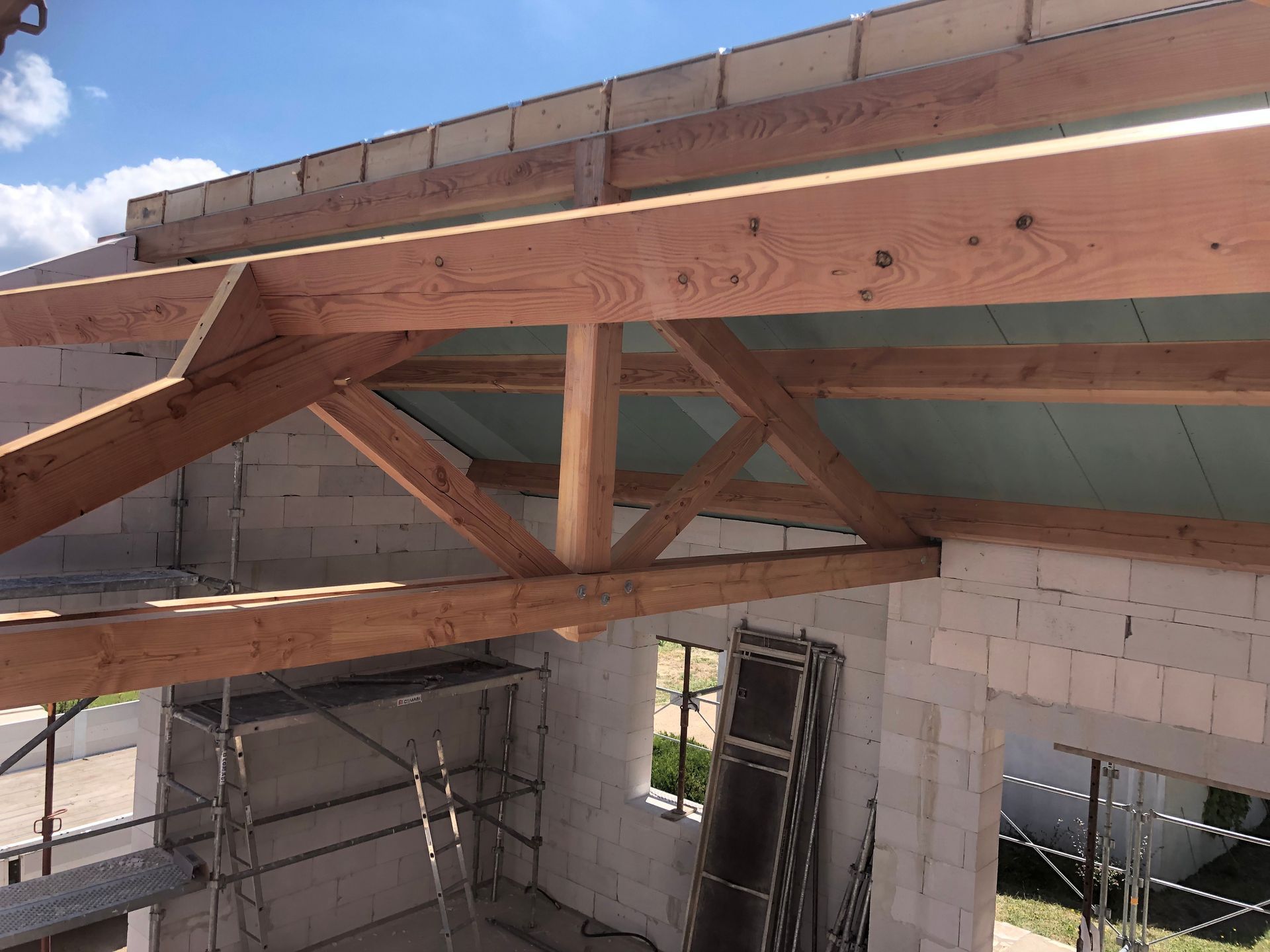 Des fermes de toit en bois sont en cours de construction sur un bâtiment en briques claires, sous un ciel bleu.
