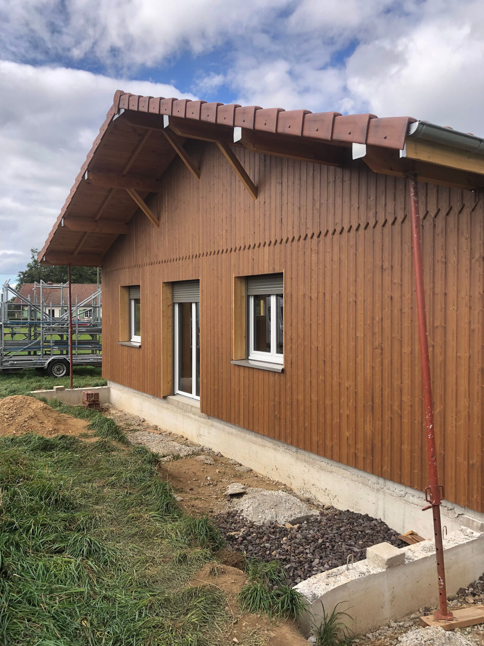 Maison en construction sur fondations en béton, revêtue de bois brun, avec fenêtres et porte blanches, sous un ciel nuageux.