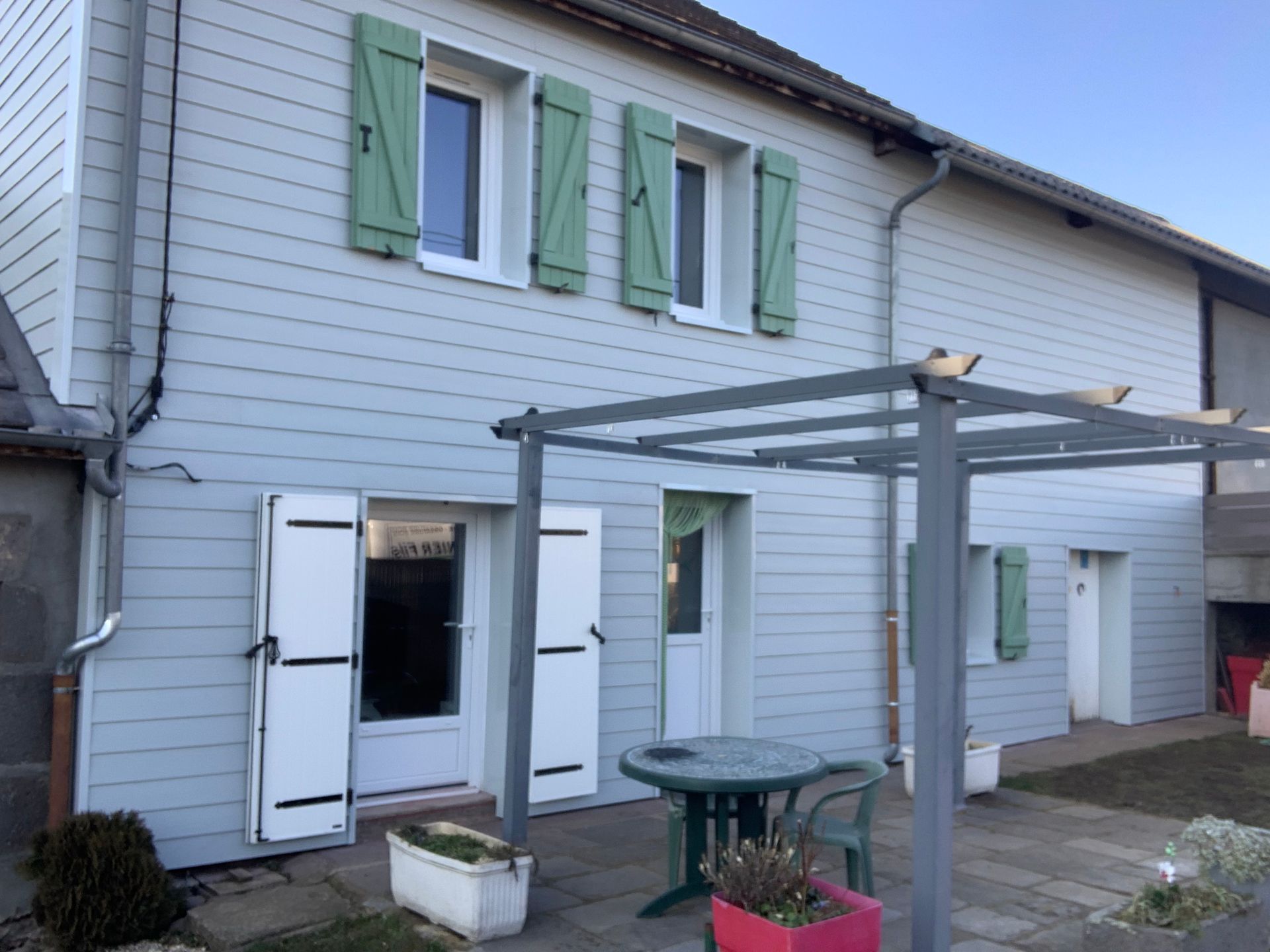 Maison bleu clair avec des volets verts, une pergola et un patio avec une table et des chaises.