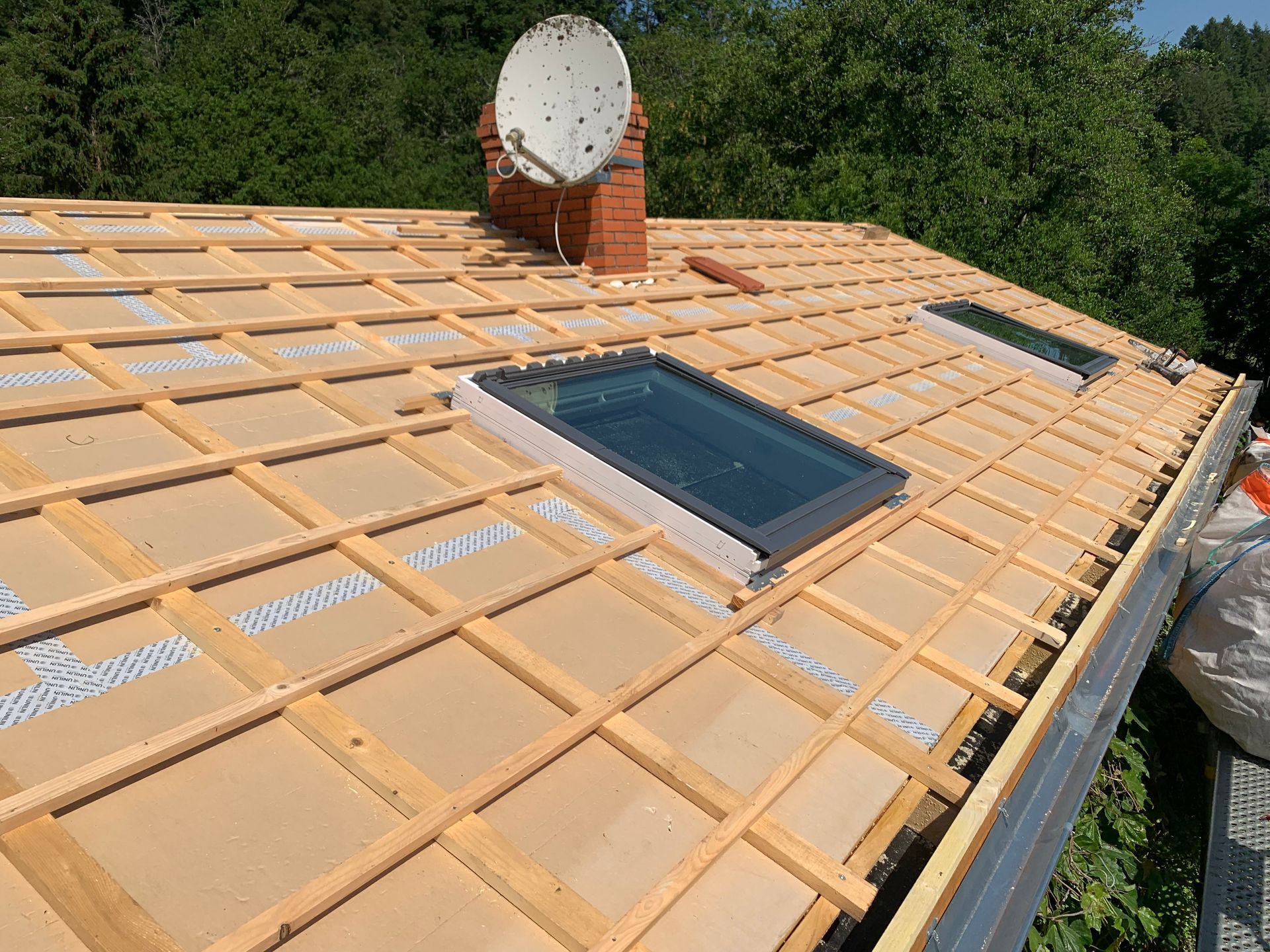 Toiture en construction avec charpente en bois, deux puits de lumière, cheminée et antenne parabolique.