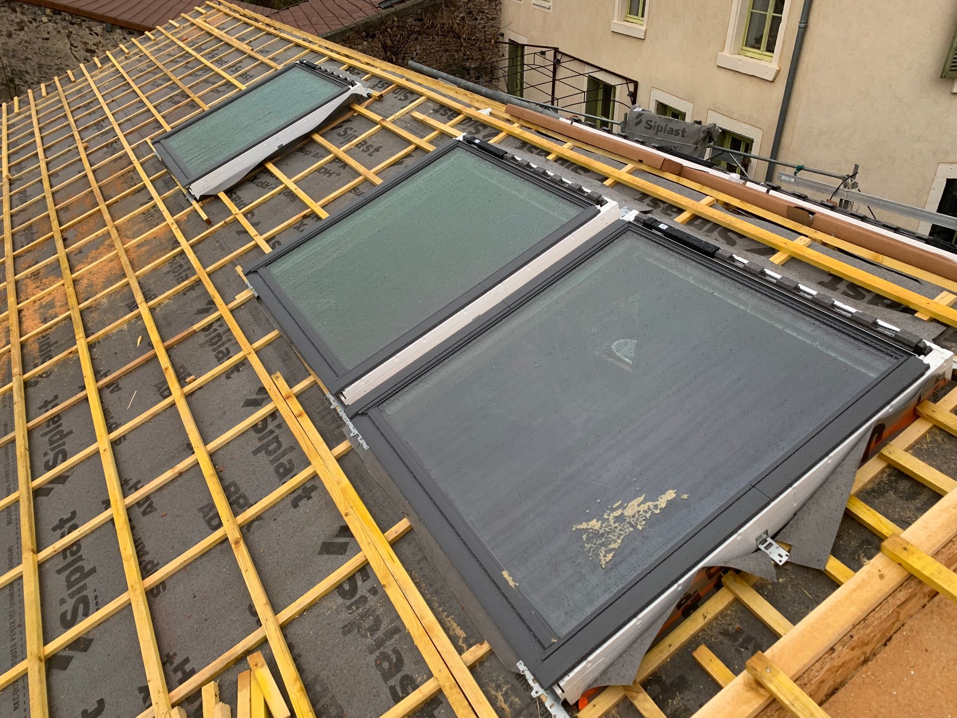 Trois puits de lumière installés sur un toit en construction ; poutres en bois et matériau de couverture noire visible.