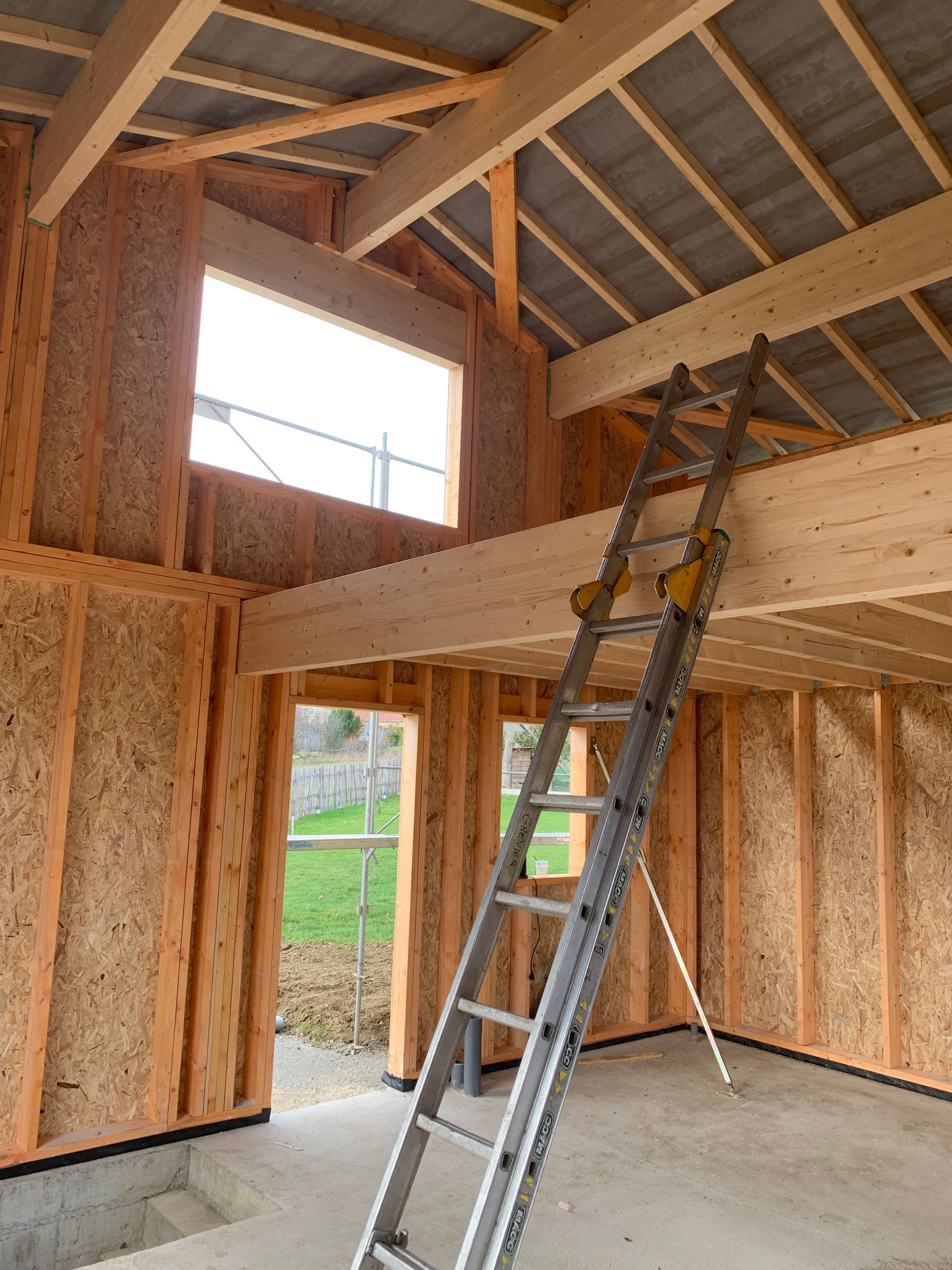 Intérieur d'un bâtiment en construction, montrant la charpente en bois, une échelle et des ouvertures de fenêtres.