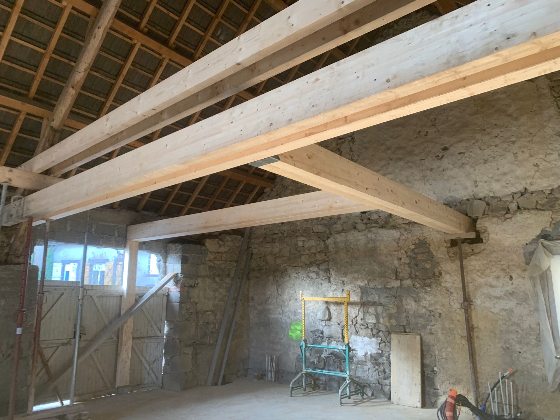 Vue intérieure d'une grange avec des poutres en bois nouvellement installées. Murs en pierre, toit visible.