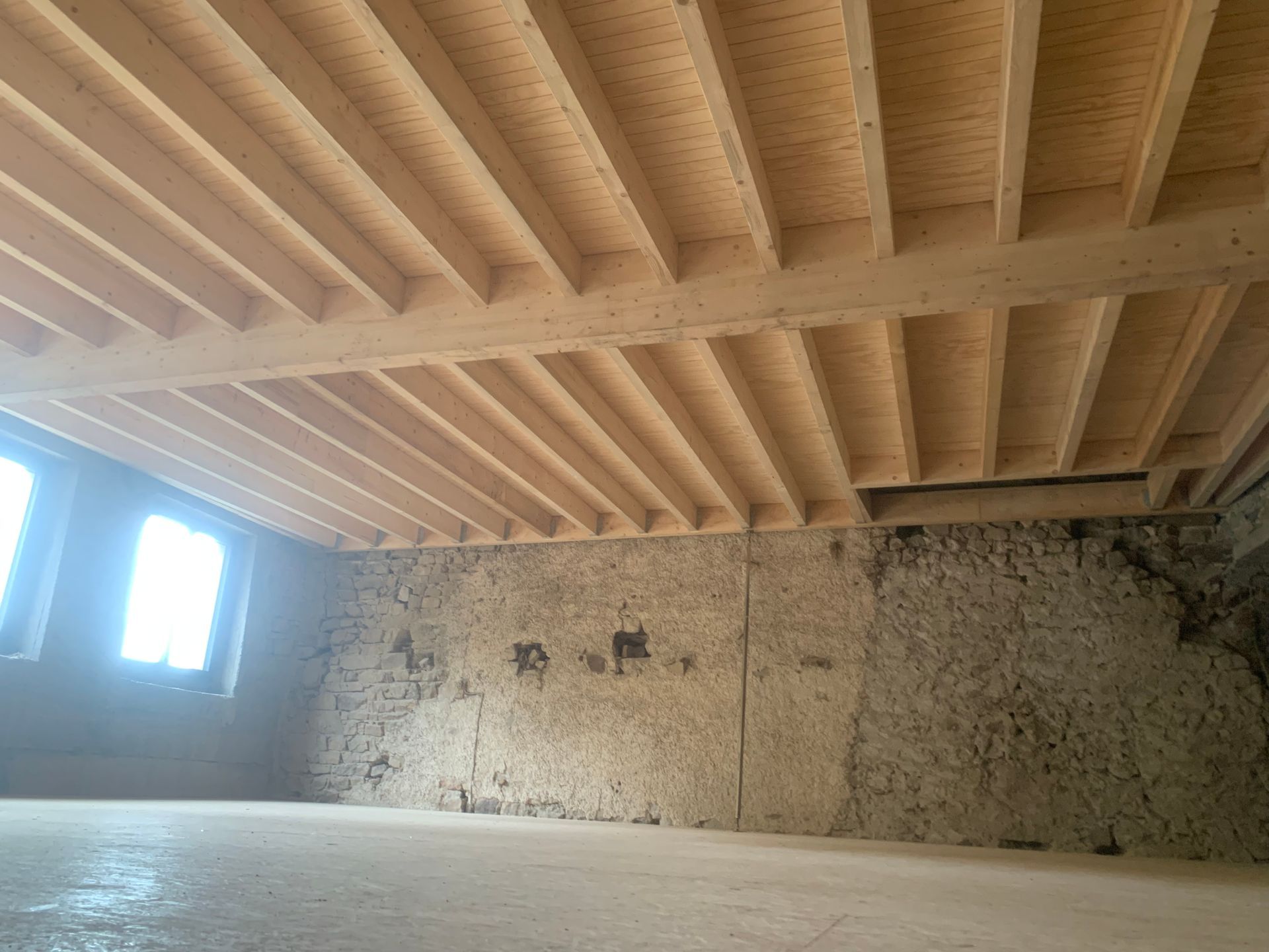 Poutres de plafond en bois et sous-plancher, au-dessus d'une pièce aux murs en pierre et comportant deux fenêtres.
