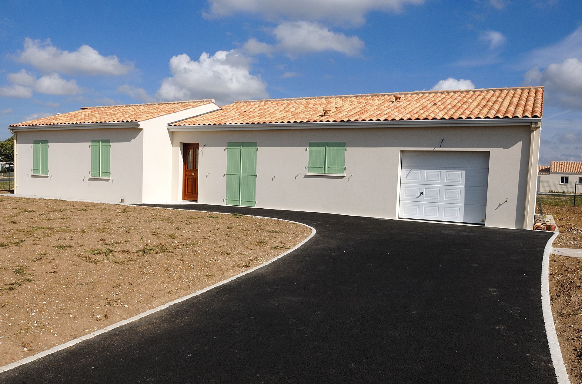 Maison de plain-pied avec façade en stuc beige, volets verts, garage et allée noire incurvée sous un ciel bleu.
