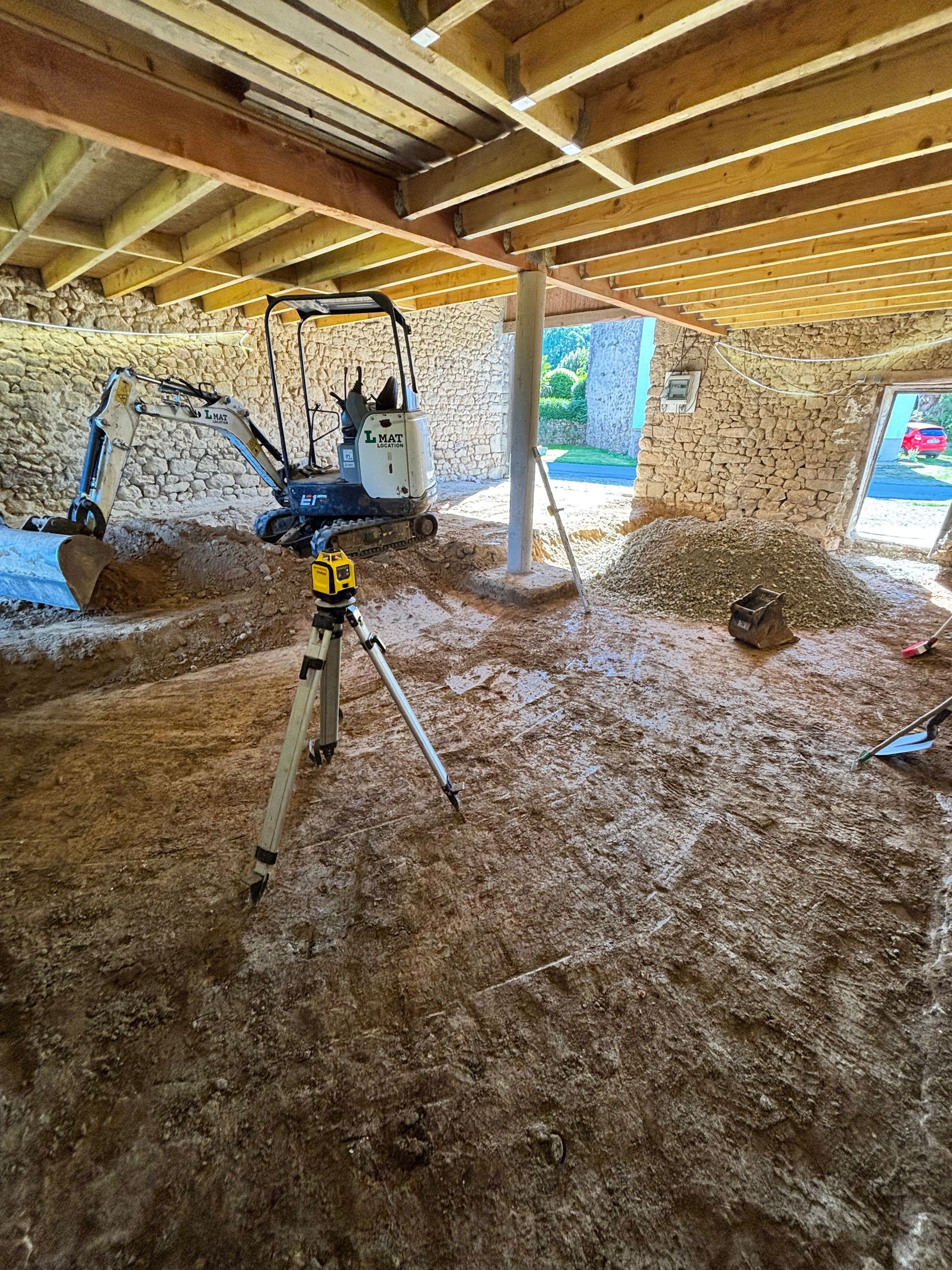 Intérieur d'une grange en construction avec charpente en bois, isolation et un outil monté sur trépied sur un sol en terre battue.