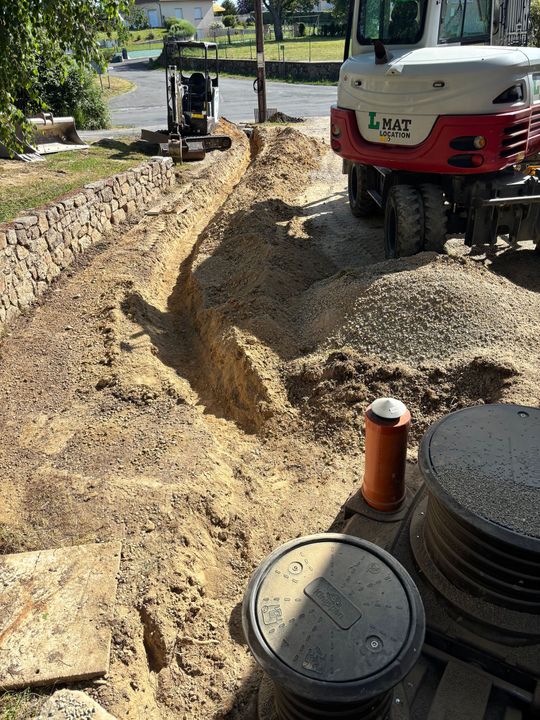 Travaux d'excavation de tranchées en bordure de route, avec une excavatrice et un camion-benne rouge déchargeant du gravier.