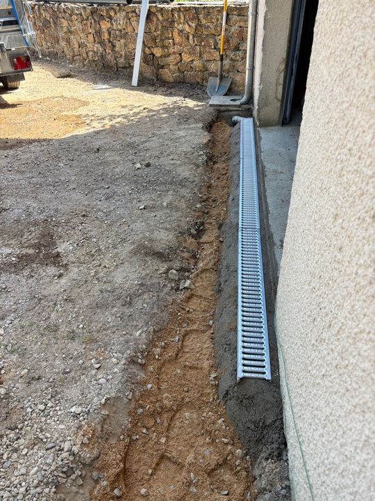 Allée de gravier longeant un mur avec un étroit caniveau de drainage et une grille métallique.