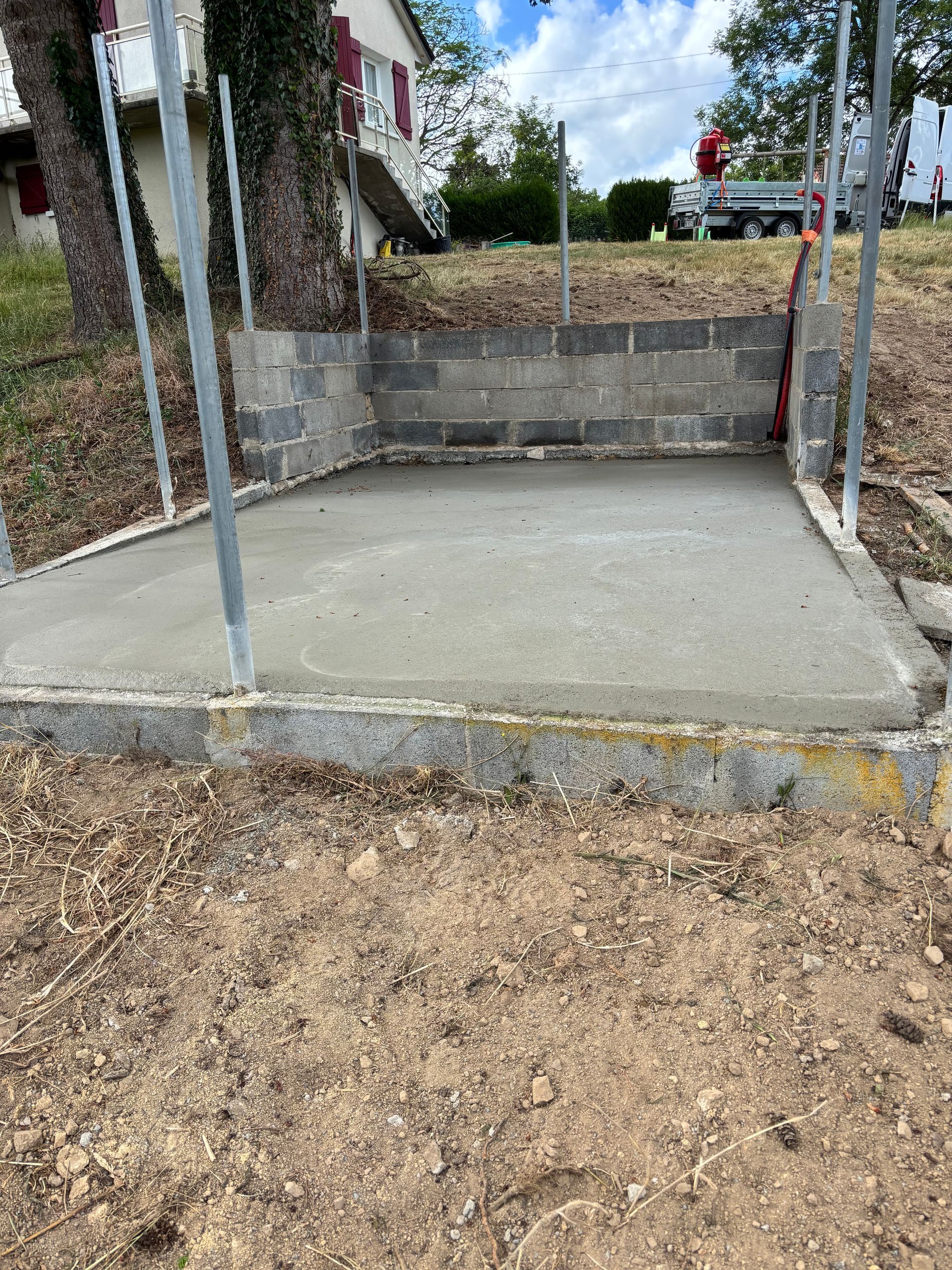 Dalle de béton dans une cour avec deux poteaux métalliques et un muret en pierre à l'arrière.