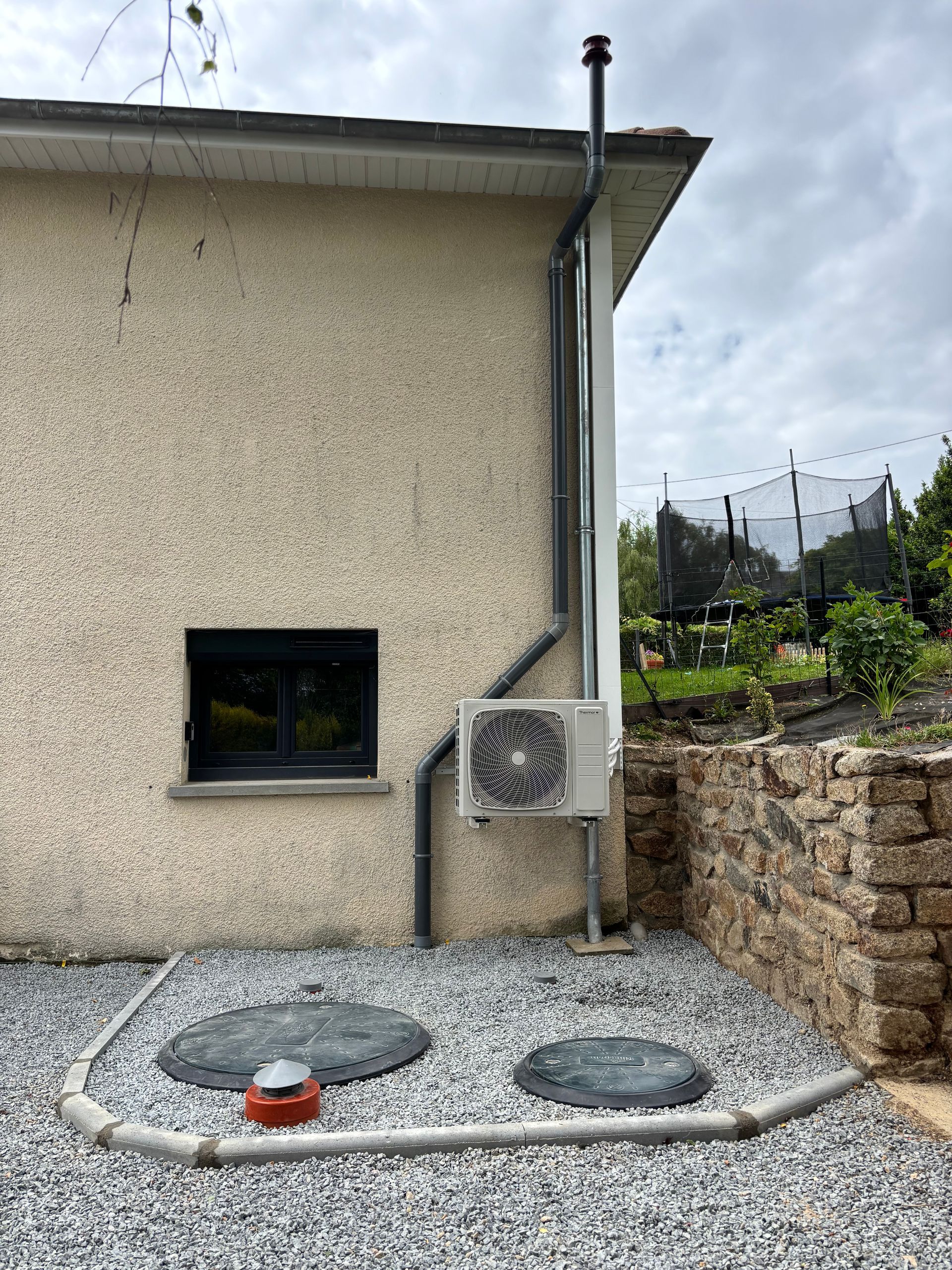 Mur extérieur avec fenêtre, descente de gouttière, unité de climatisation et deux plaques d'égout, à côté d'une bande de gravier et d'un mur de soutènement en pierre.