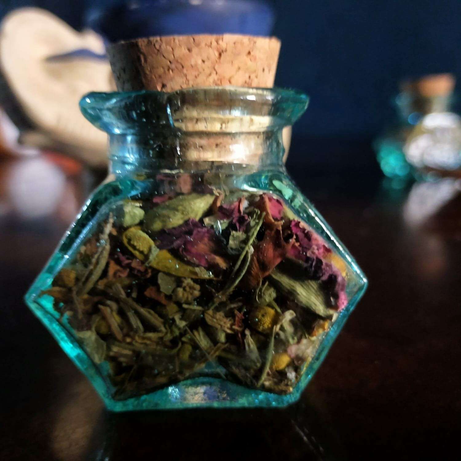 Un pequeño frasco de vidrio color verde azulado con forma de estrella y tapa de corcho, lleno de pétalos de flores secas.